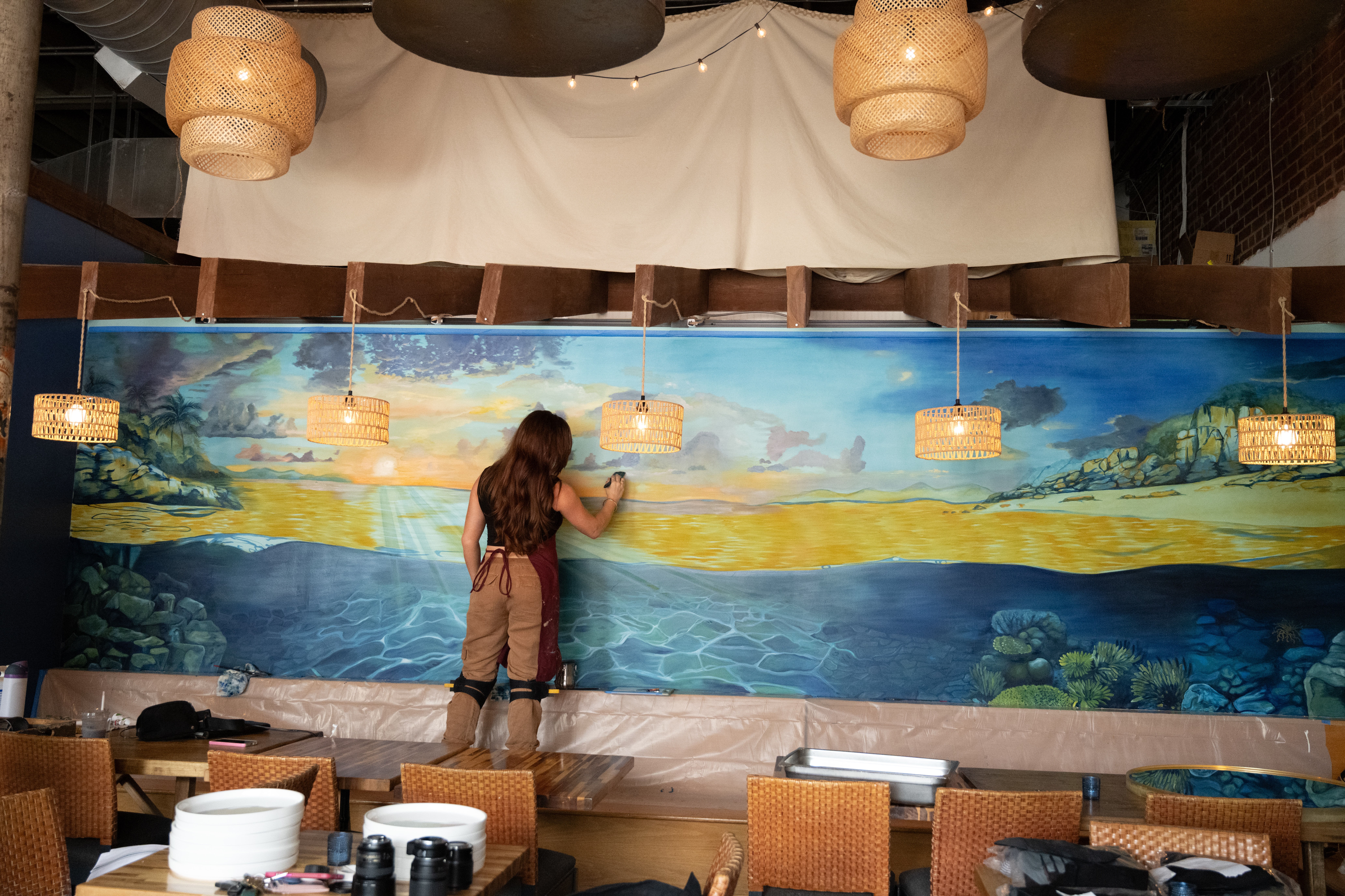 Person painting a large landscape mural on a wall, with tables and hanging lights in a room.