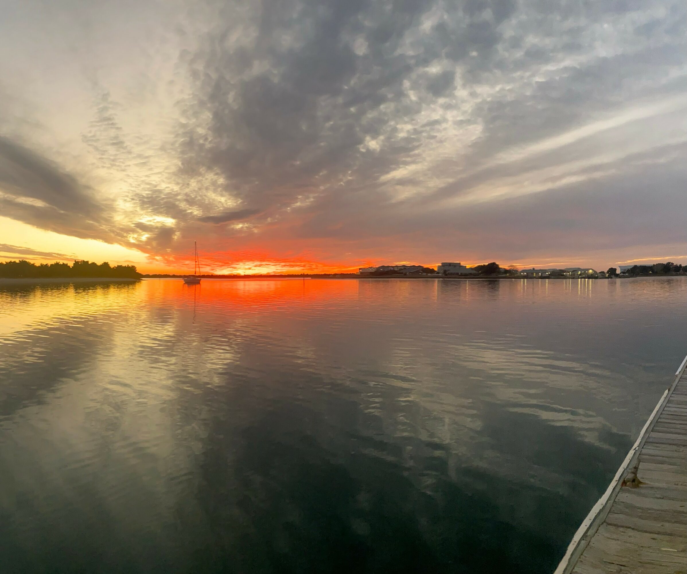Sunset over a calm body of water with a dock on the right, clouds in the sky, and reflections on the water surface.