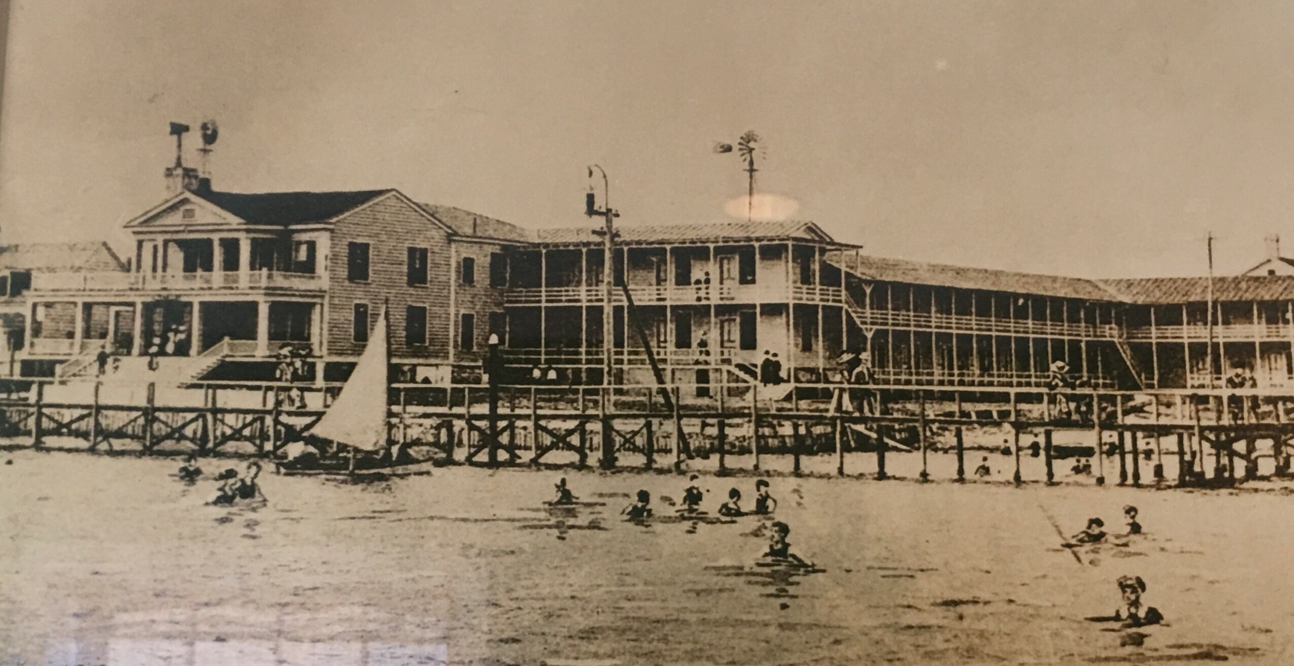 Historical photo of a large building by the water with people swimming and boats nearby.