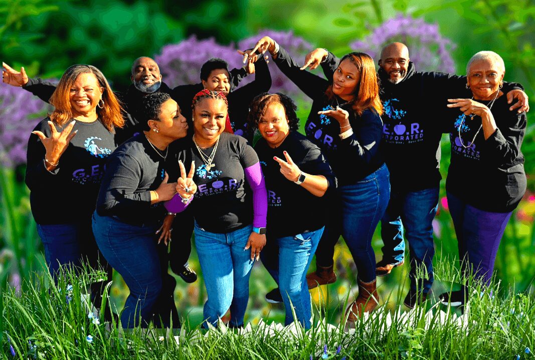 Group of nine people outdoors, smiling, holding hands, surrounded by green grass and purple flowers, bright and cheerful scene.
