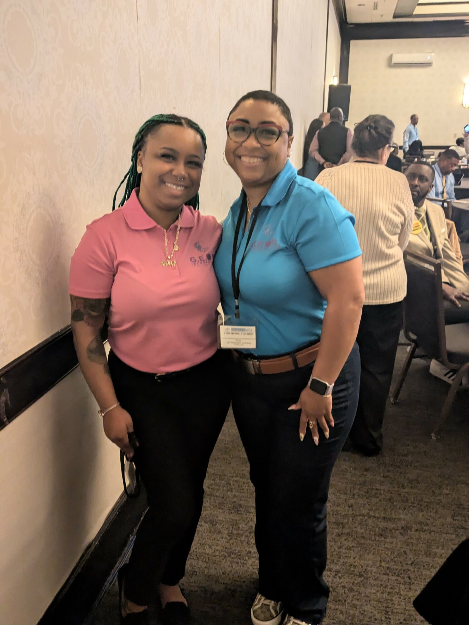 Two women standing together, smiling, in a conference room with other people in the background.