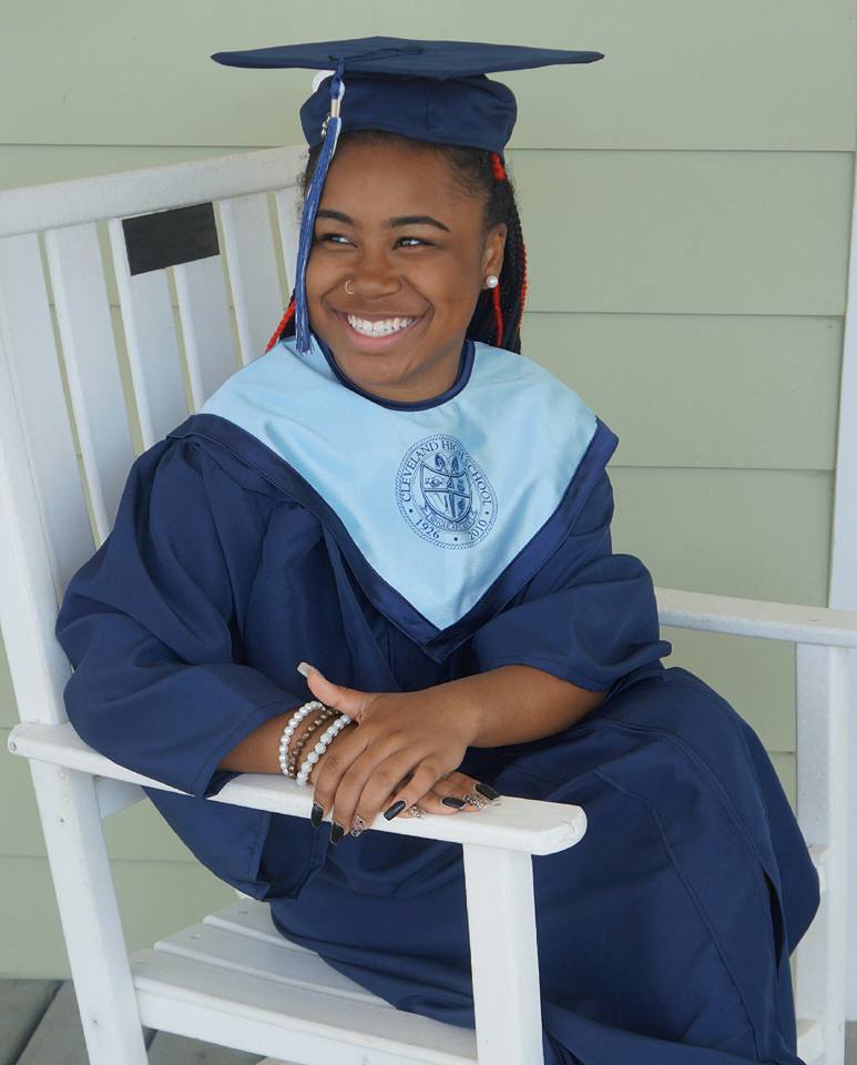 Young girl in graduation gown and cap, smiling, sitting on a white chair, holding her hands together, against a light green wall.