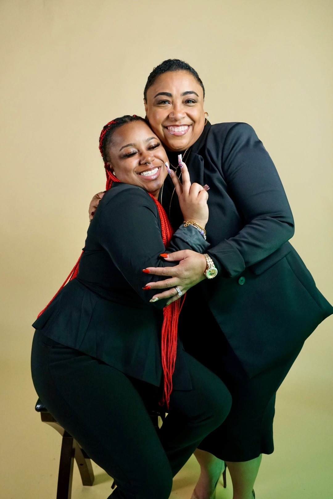 Two women smiling, hugging, and posing together against a plain background.