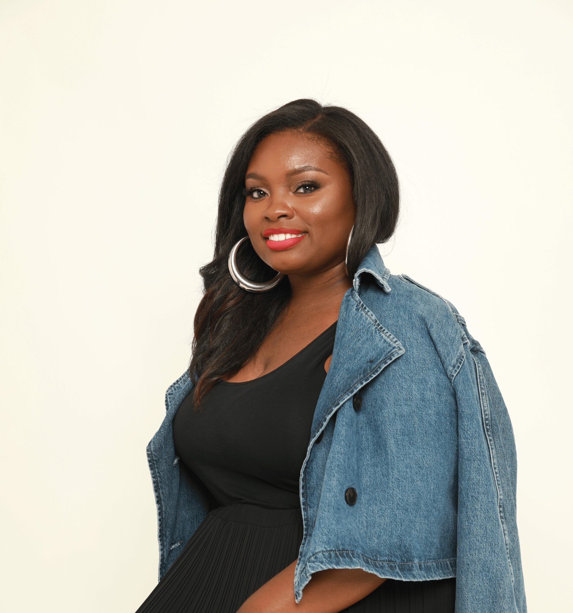 Woman with long dark hair, wearing hoop earrings, a black top, and a denim jacket, smiling against a plain background.