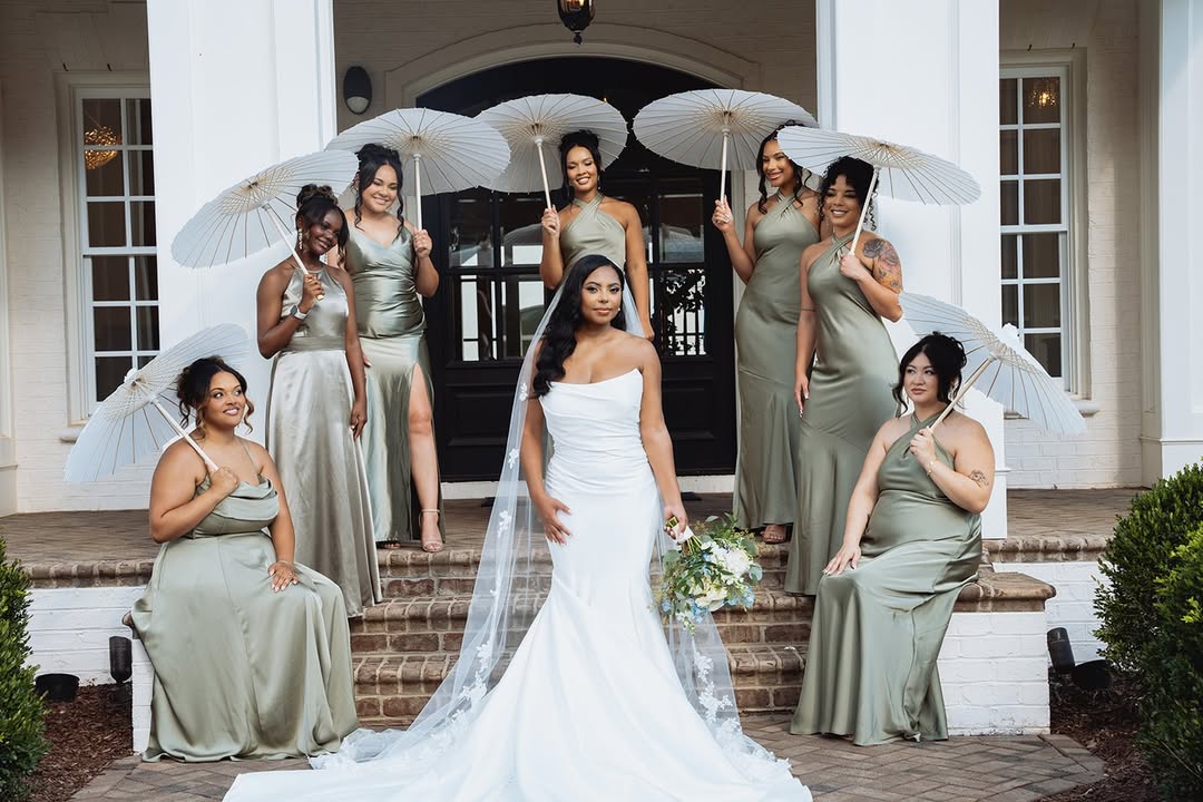 Bride in white gown with veil holding bouquet, surrounded by bridesmaids in matching dresses holding umbrellas on steps outside a building.