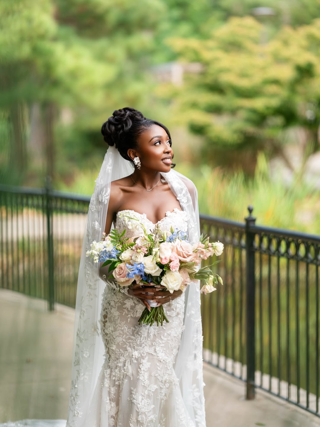 Bride in wedding dress holding bouquet outdoors, smiling, with trees and a fence in background.