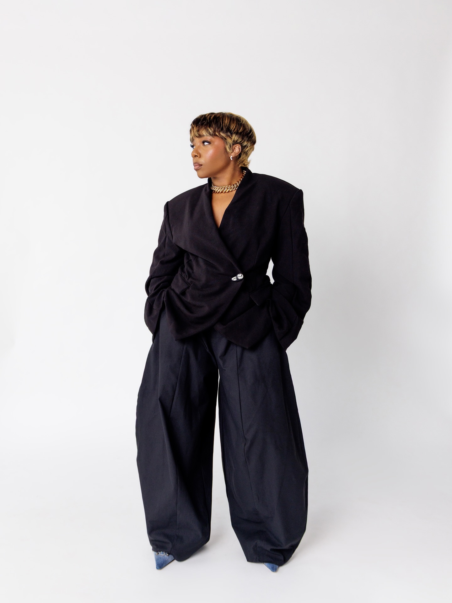 Woman with short curly hair wearing a black blazer and wide black pants, standing against a white background.
