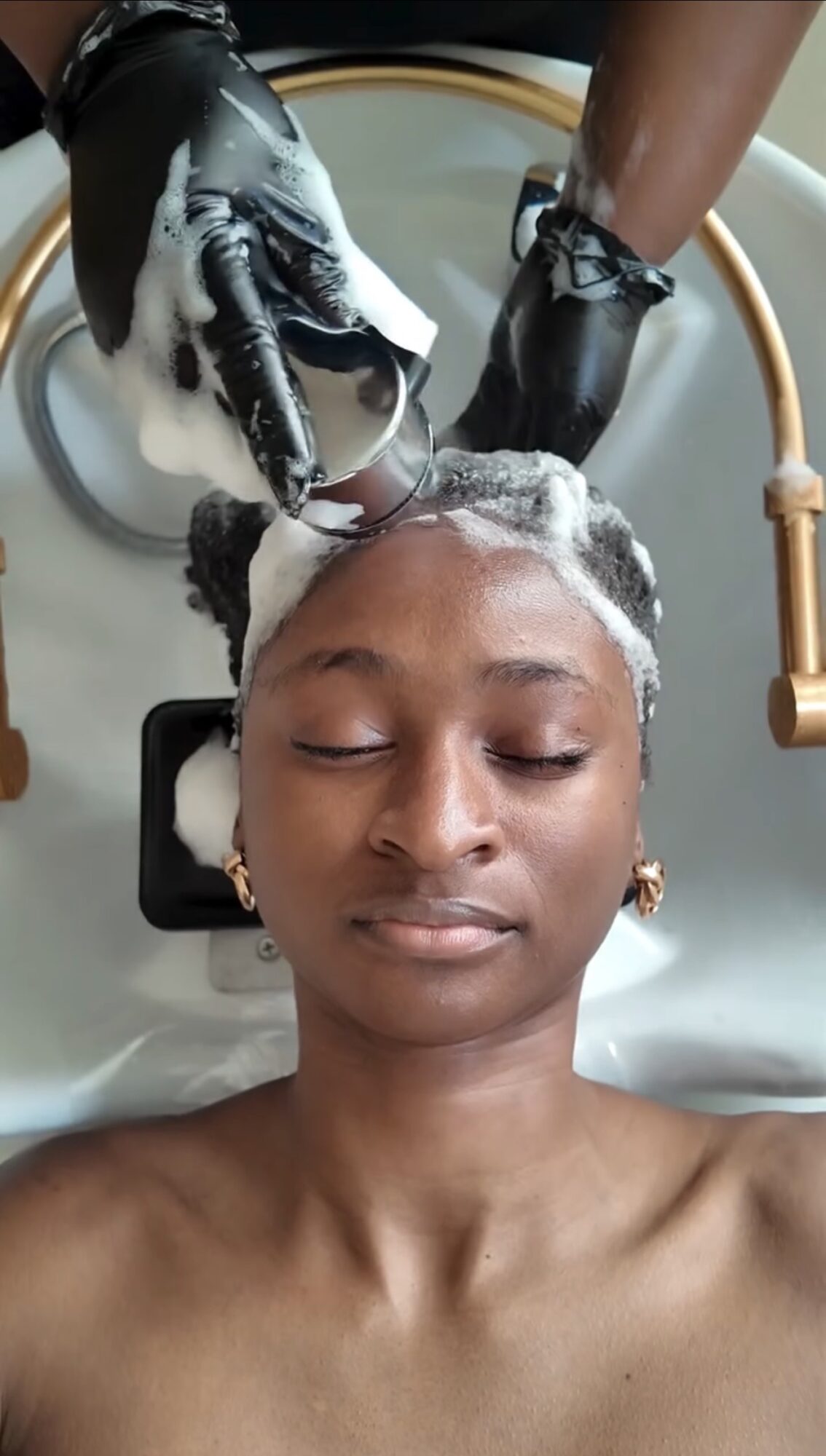 Person with closed eyes receiving a hair wash, with soap lather on hair, in a salon setting.