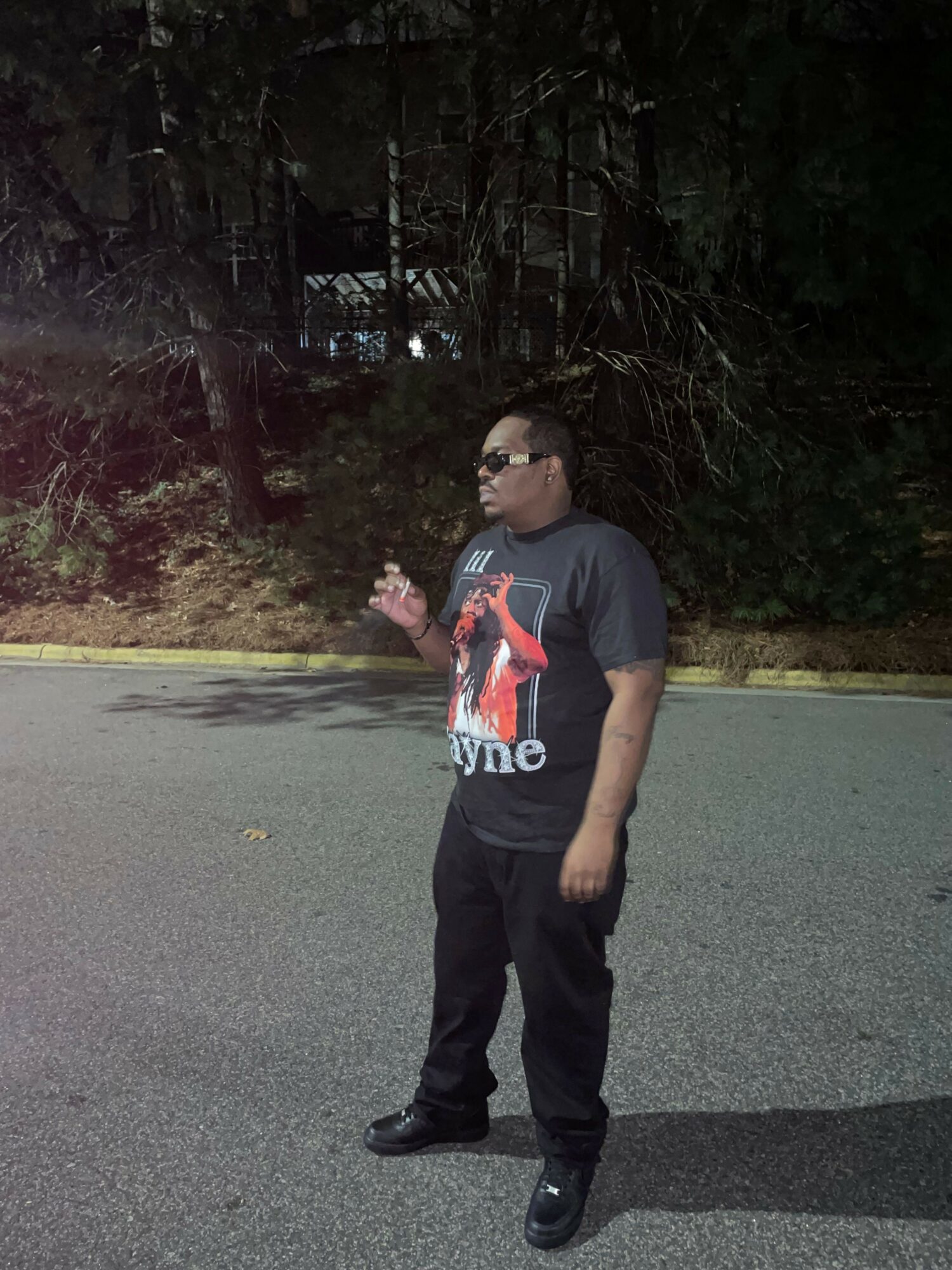 Person standing on a paved surface with trees in the background, wearing a dark t-shirt and pants, holding a phone.