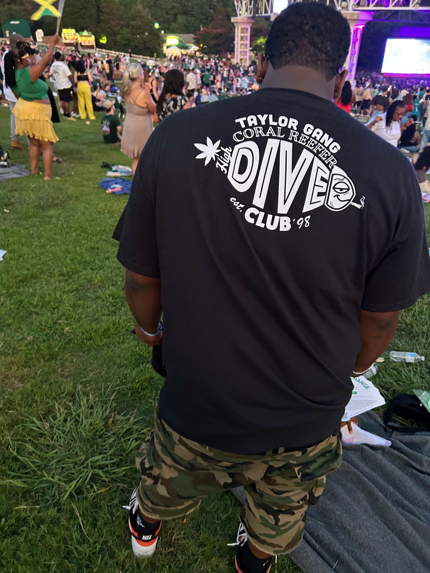 Person wearing a black hoodie with white text, camouflage pants, and sneakers, sitting on grass at an outdoor event.