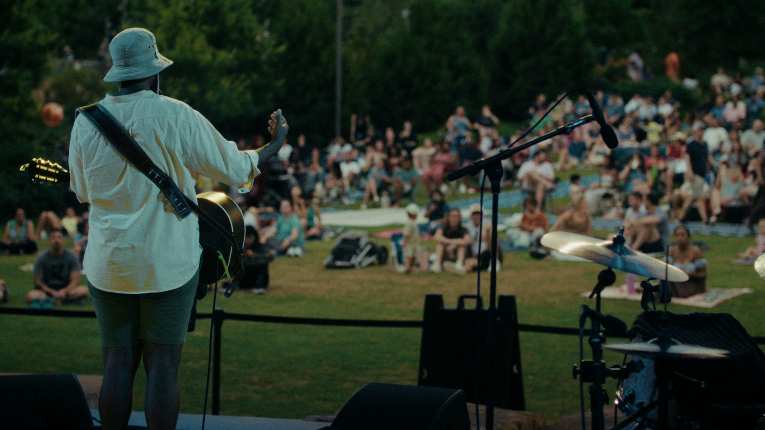 Person playing guitar on stage with a large crowd sitting on grass in park, trees in background.