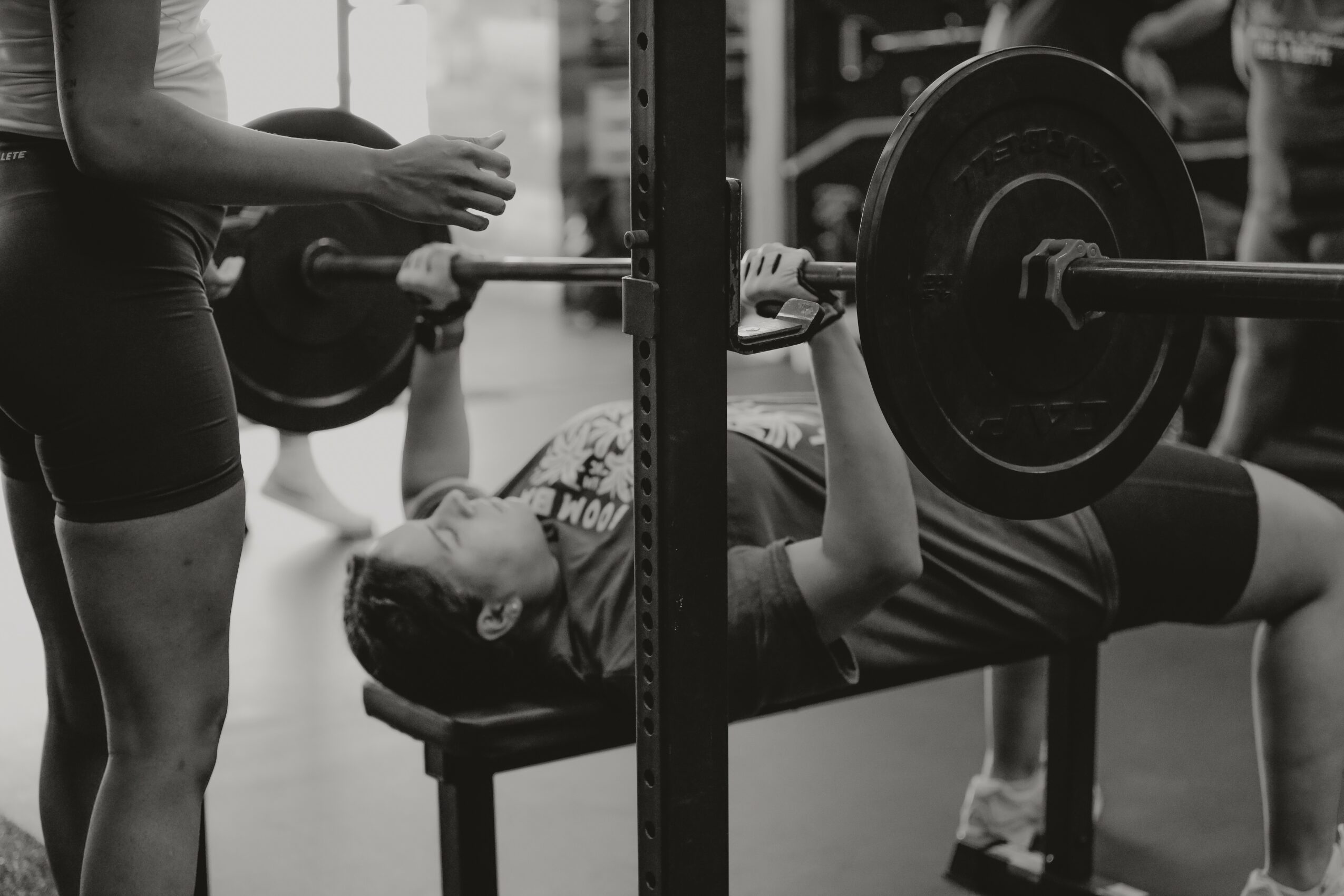 Person lying on bench pressing weights with a spotter nearby, gym setting, black and white photo.