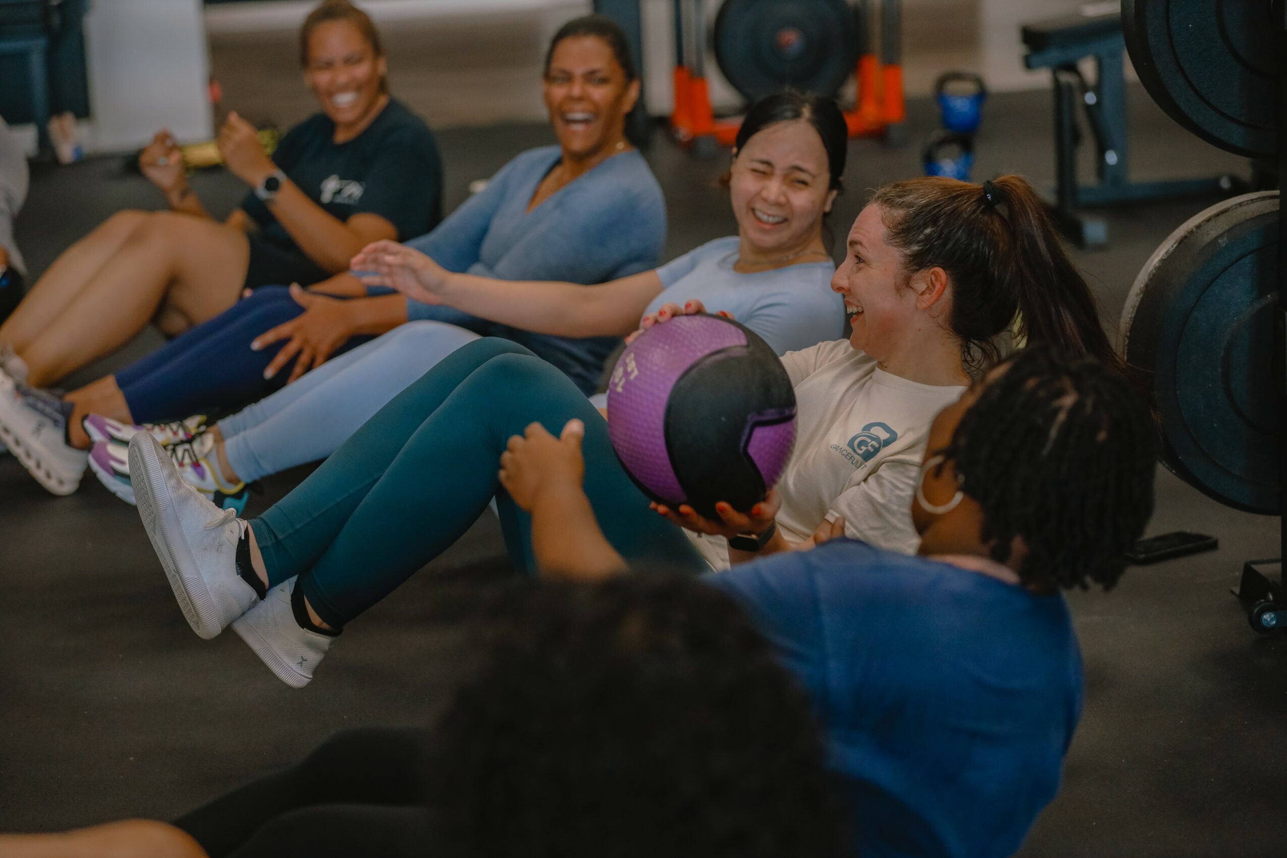 Group of women sitting on the floor in a gym, smiling, with one holding a purple exercise ball.