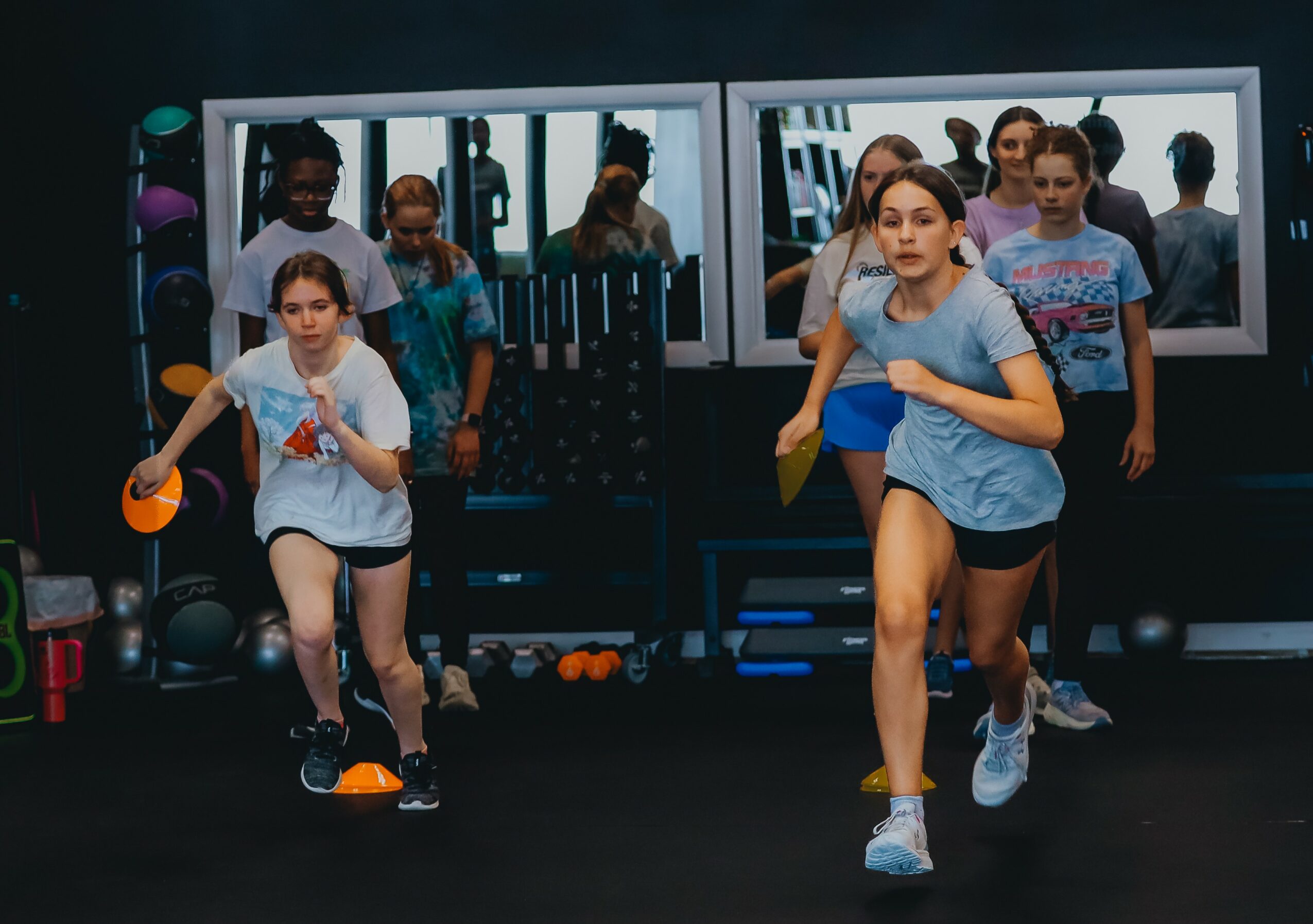 Group of children running in a gym with mirrors and exercise equipment in the background.