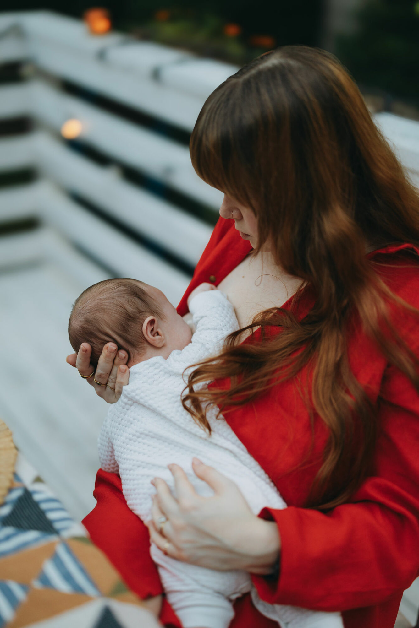 Woman in red holding a baby in white clothing outdoors, with a white fence in the background.