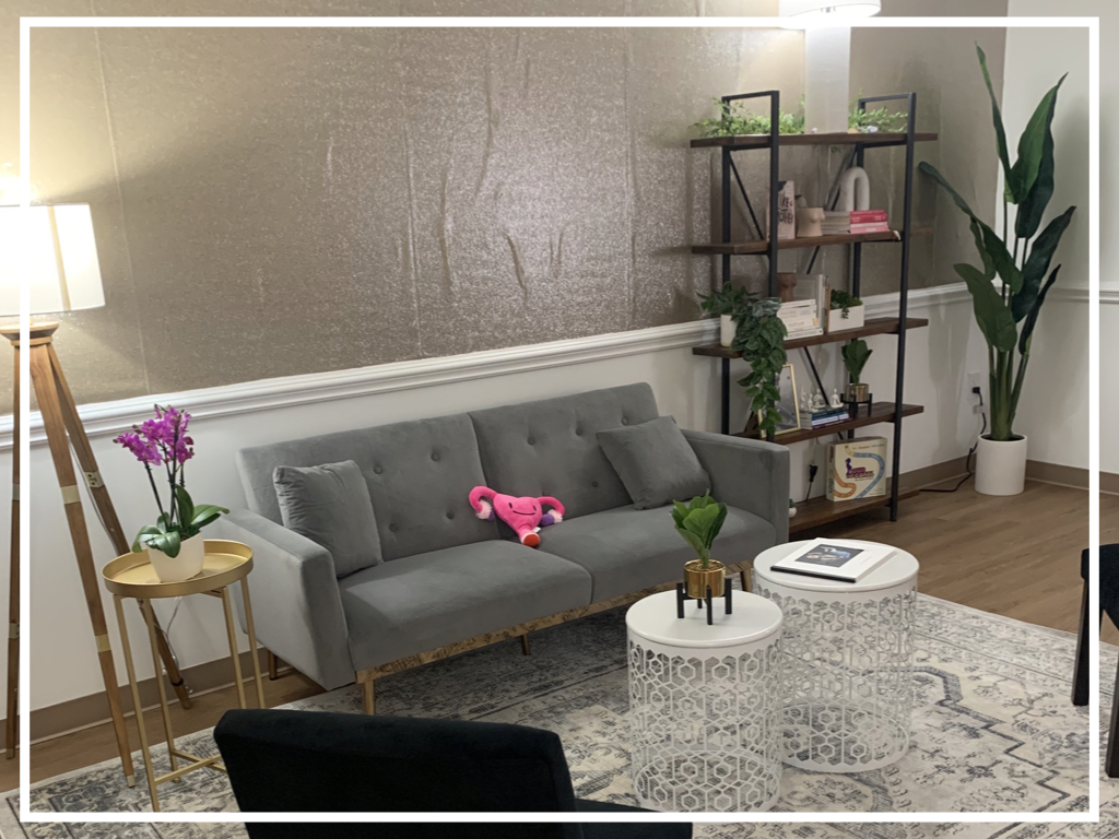 Living room with gray sofa, side table with purple flowers, bookshelf, and large plant, viewed from the side.