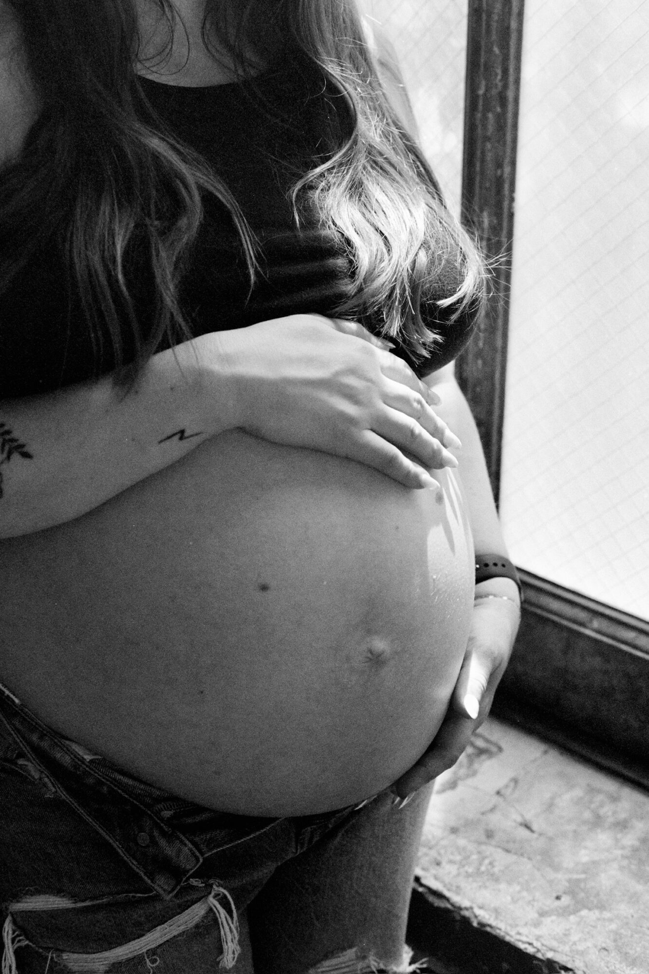 Pregnant person holding belly near window, with hand resting on abdomen, in black and white.