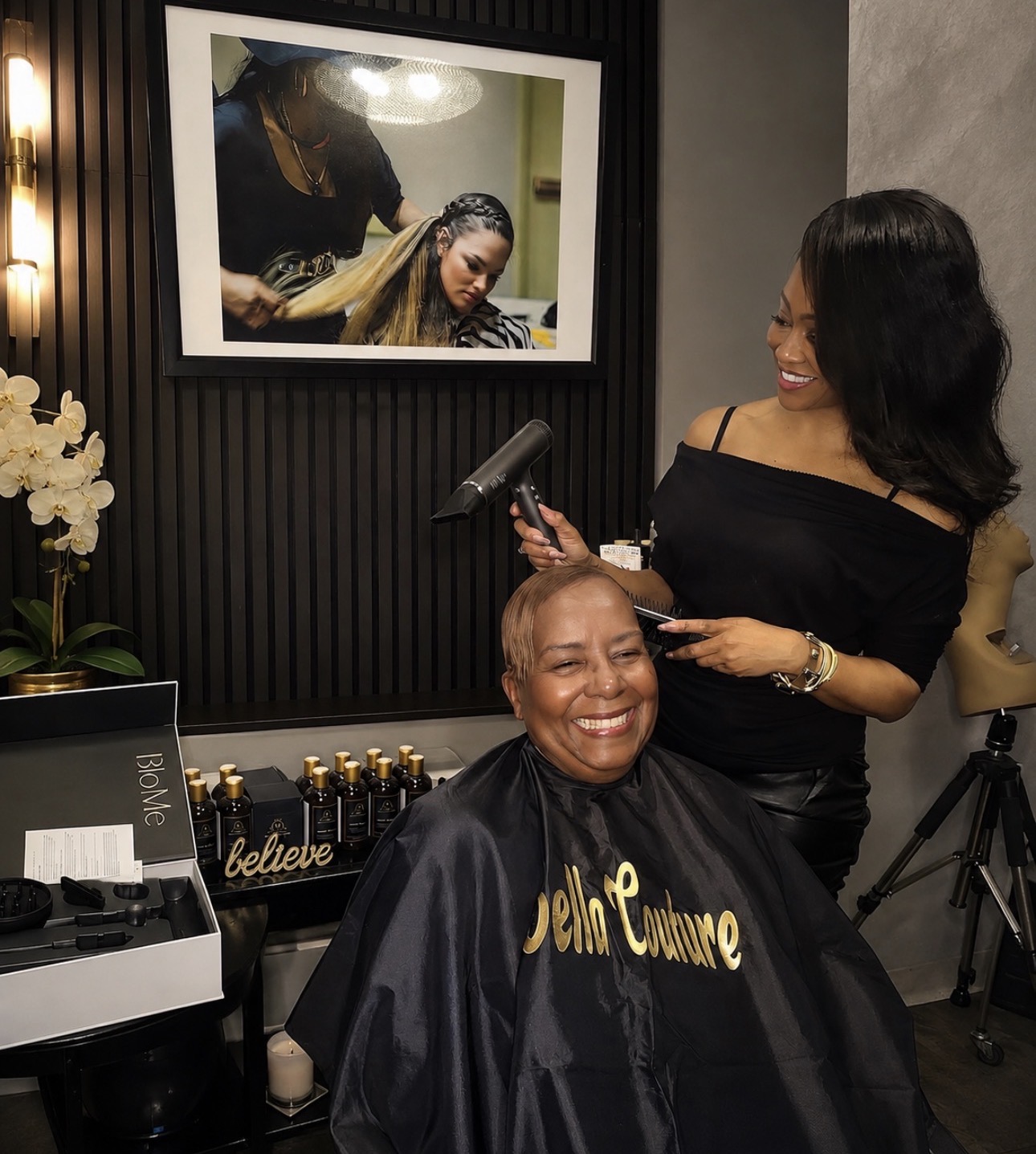 A woman with short hair smiling at a salon, a stylist with long hair holding a hairbrush, mirror reflection shows another woman. Word 'believe' visible.