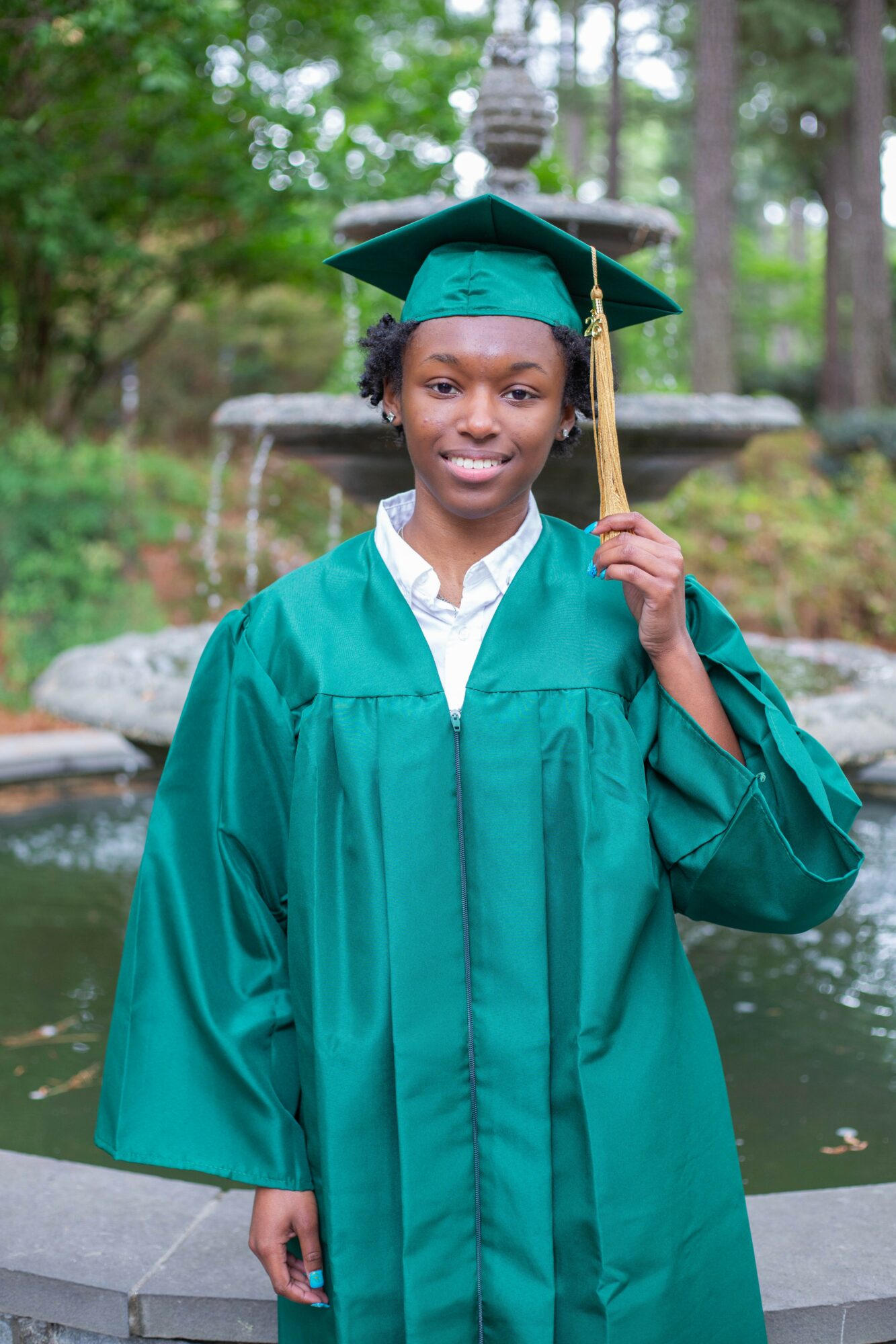 Young woman in green graduation gown and cap, holding tassel, standing outdoors near fountain with trees in background.