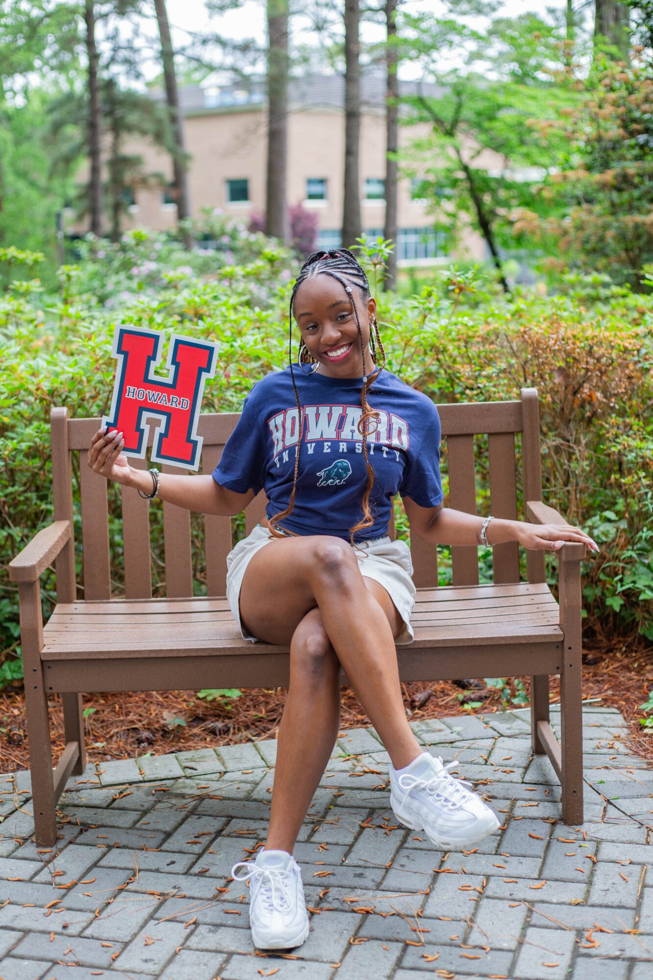 Young woman sitting on a park bench holding a University of Houston sign, smiling, surrounded by trees and greenery.