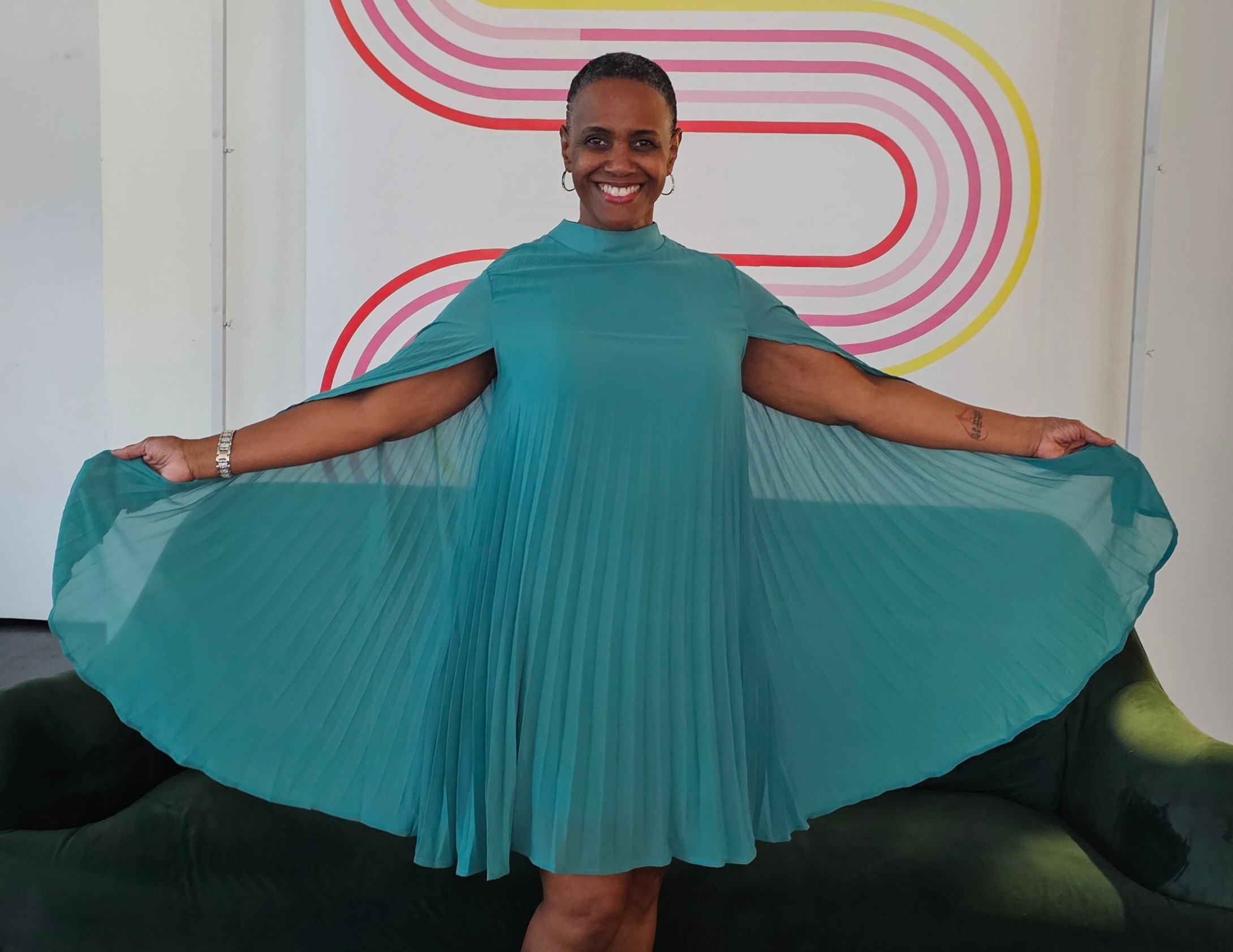 Smiling woman in a teal pleated dress holding out the sides, standing in front of a colorful background.
