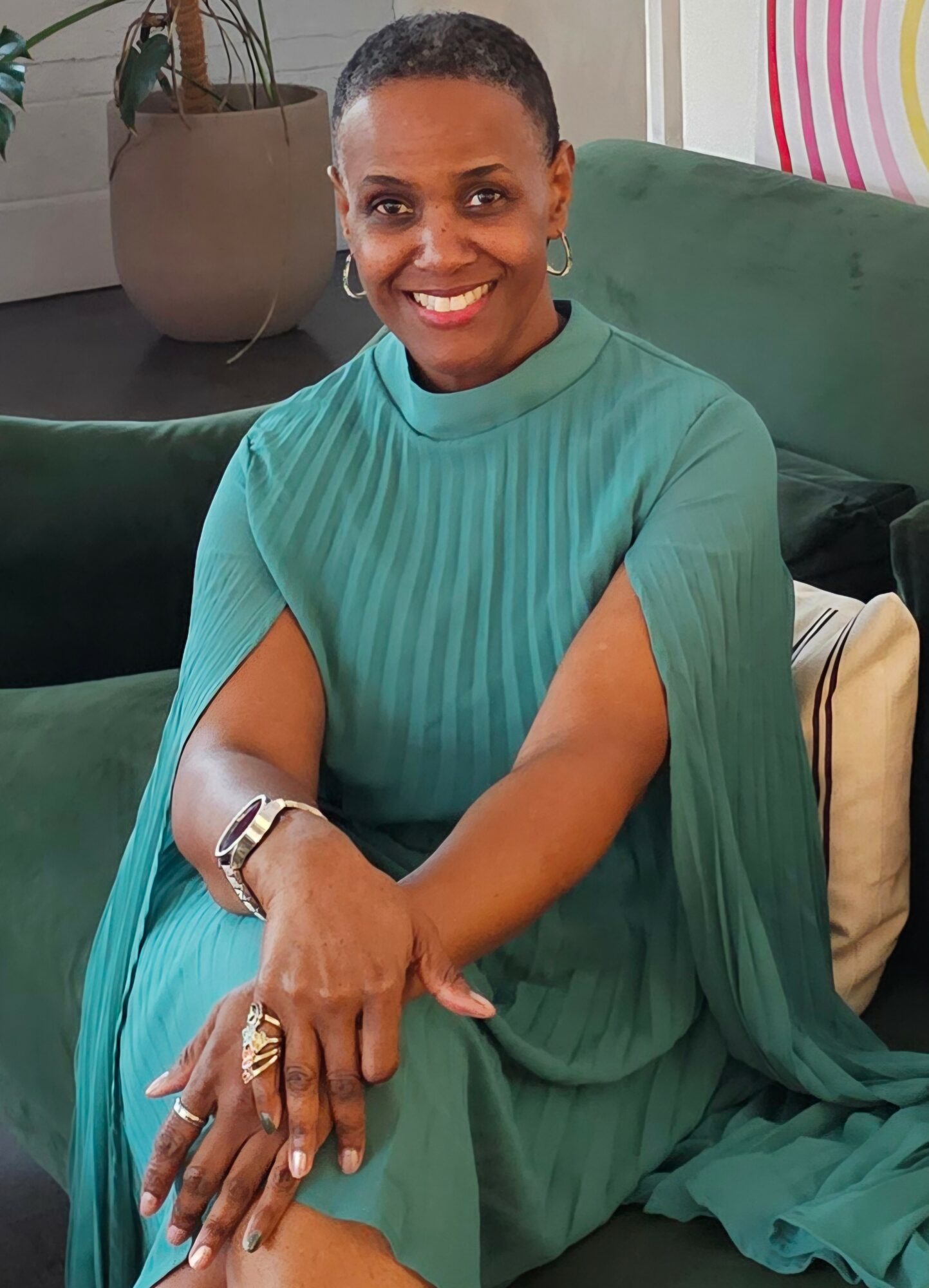Smiling woman with short hair wearing a teal dress, sitting on a green sofa with a beige pillow, in a room with a plant and artwork.