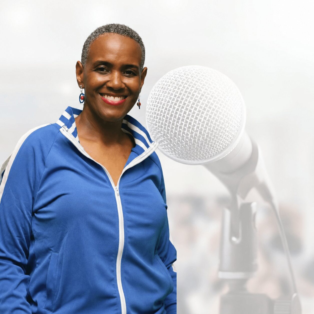 Smiling woman with short hair wearing a blue jacket, standing outdoors near a microphone, with a blurred background.