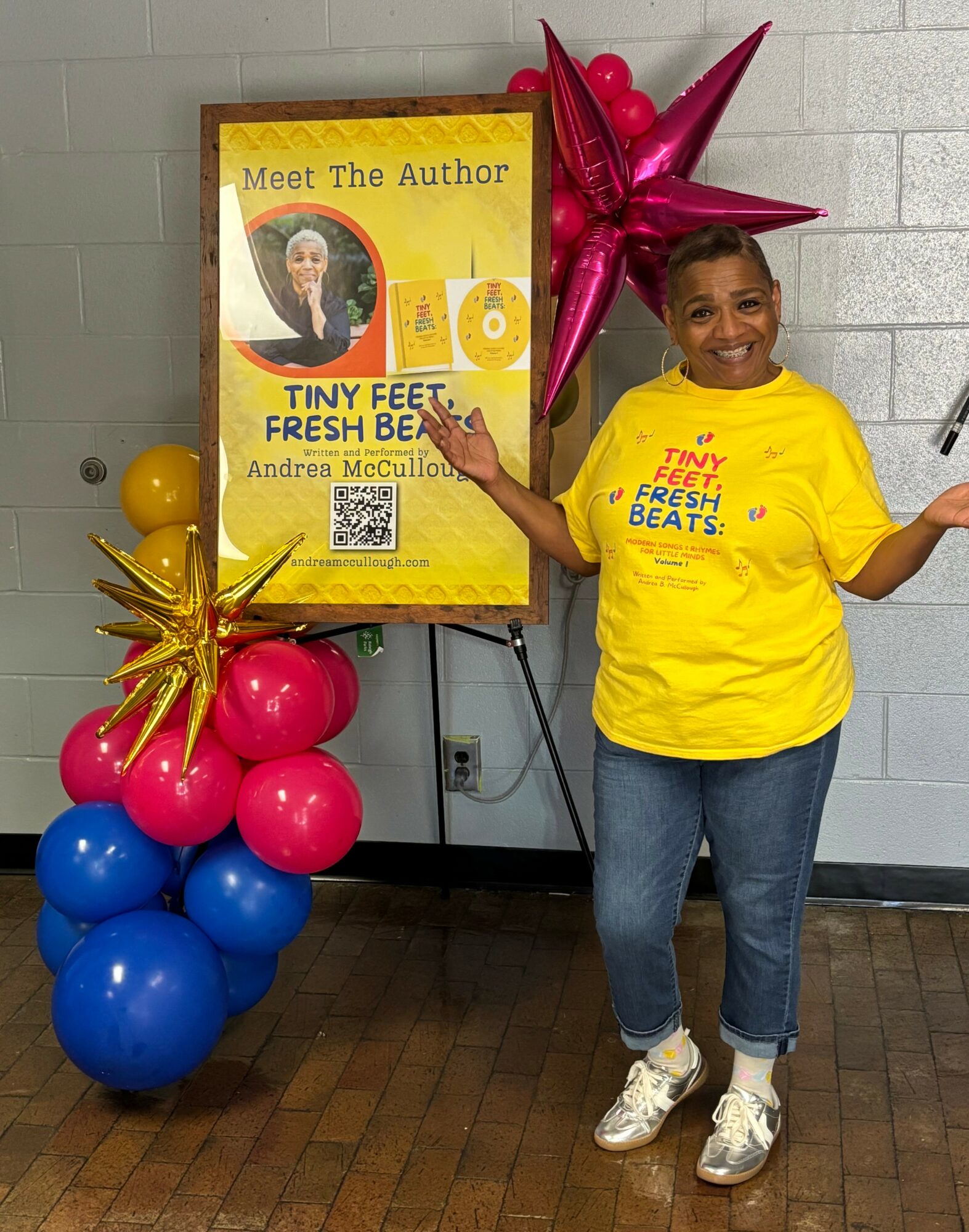 Woman standing next to a book display with balloons and star-shaped decorations, smiling and gesturing with her hands.