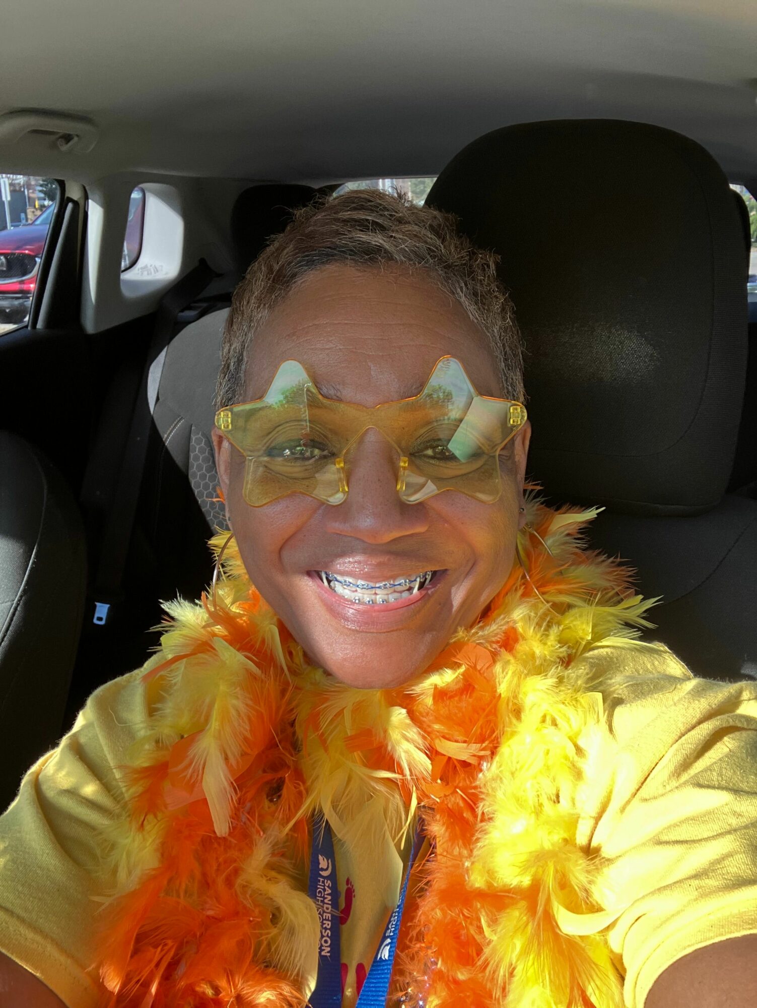 Person smiling in a costume with yellow and orange feathers, wearing glasses, inside a vehicle.