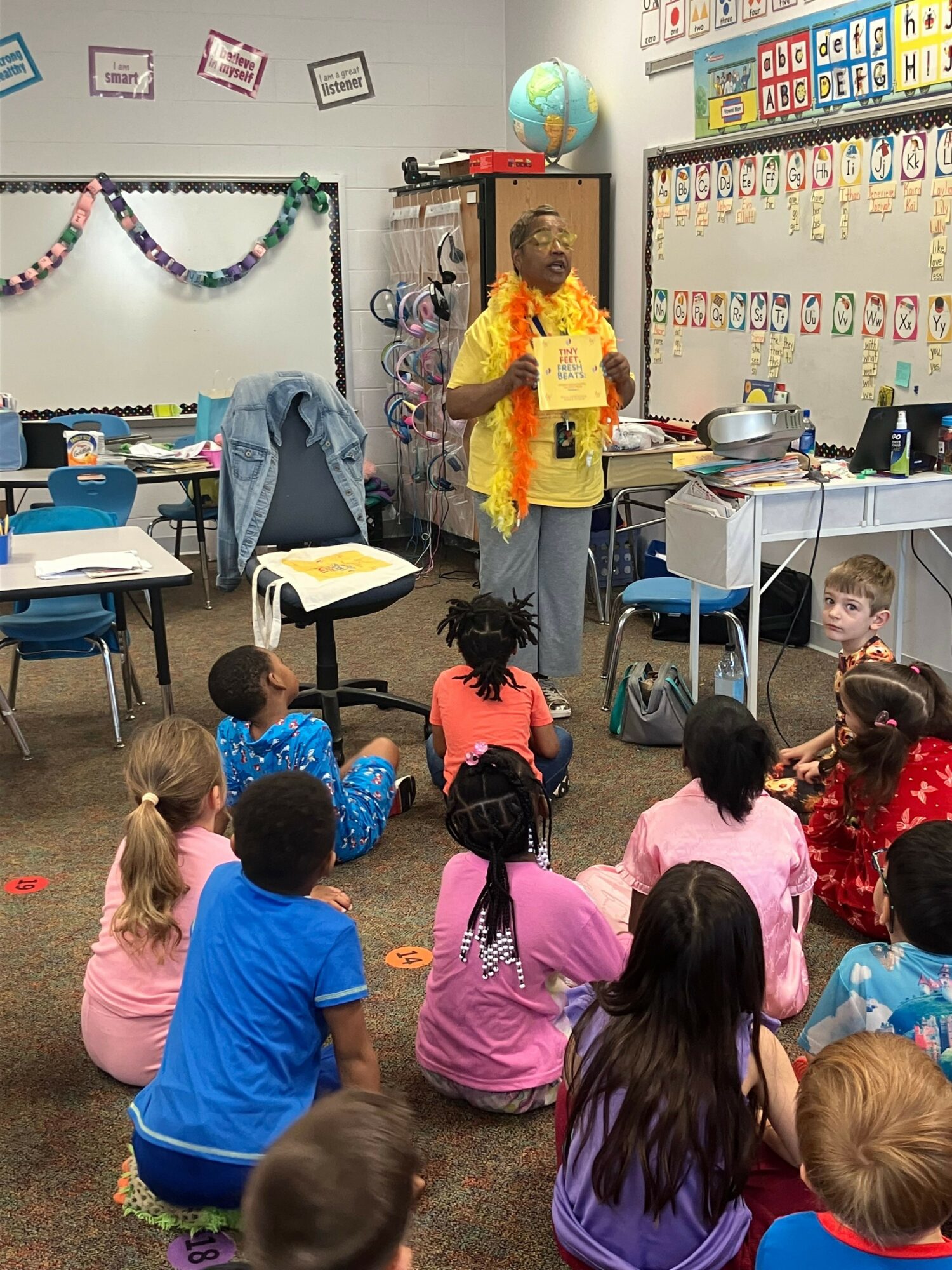 Teacher in yellow jacket stands in front of children in a classroom, with educational posters and decorations on the wall.