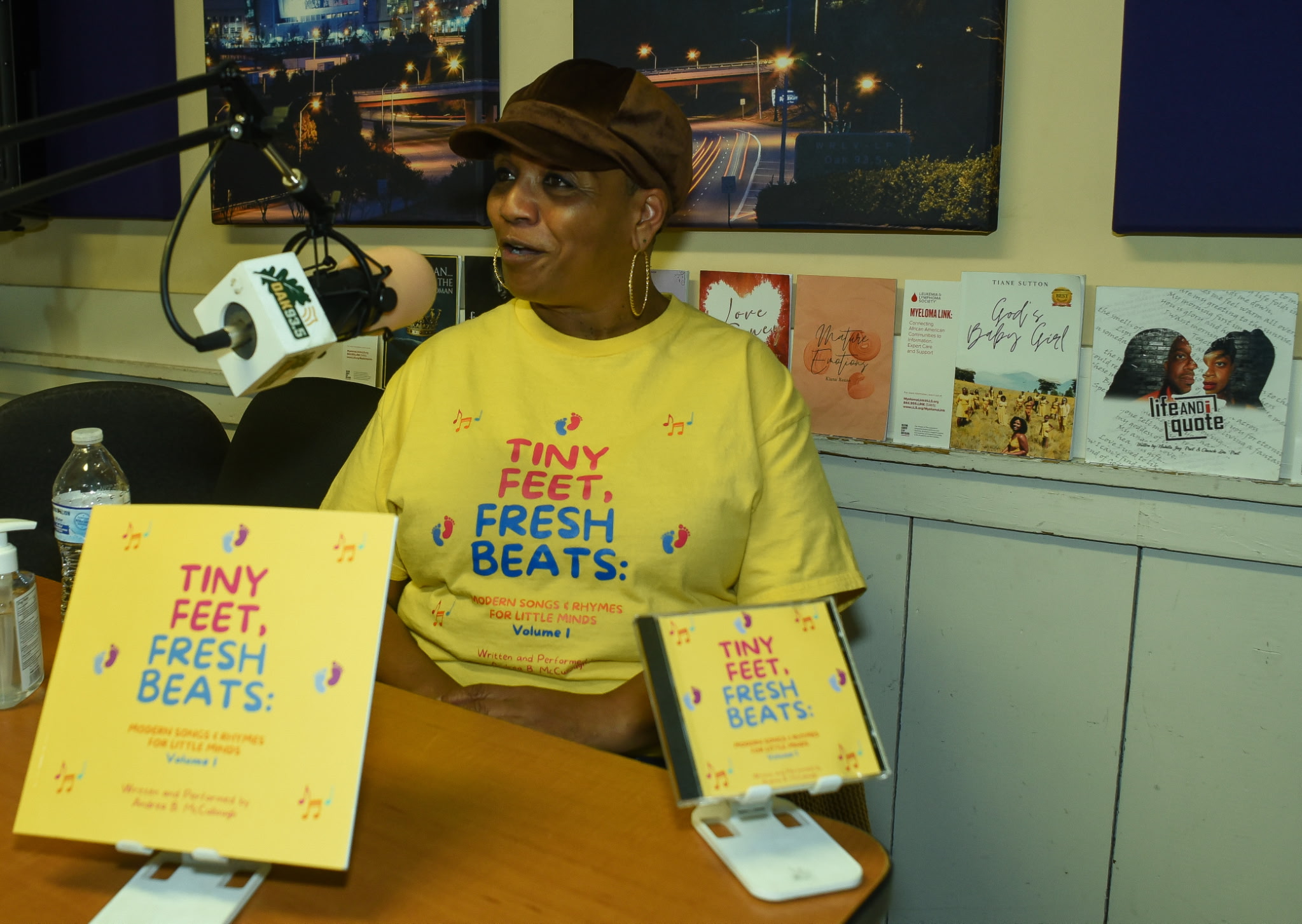 Woman wearing a yellow T-shirt and hat sitting at a table with books and a microphone in a room with artwork on the wall.