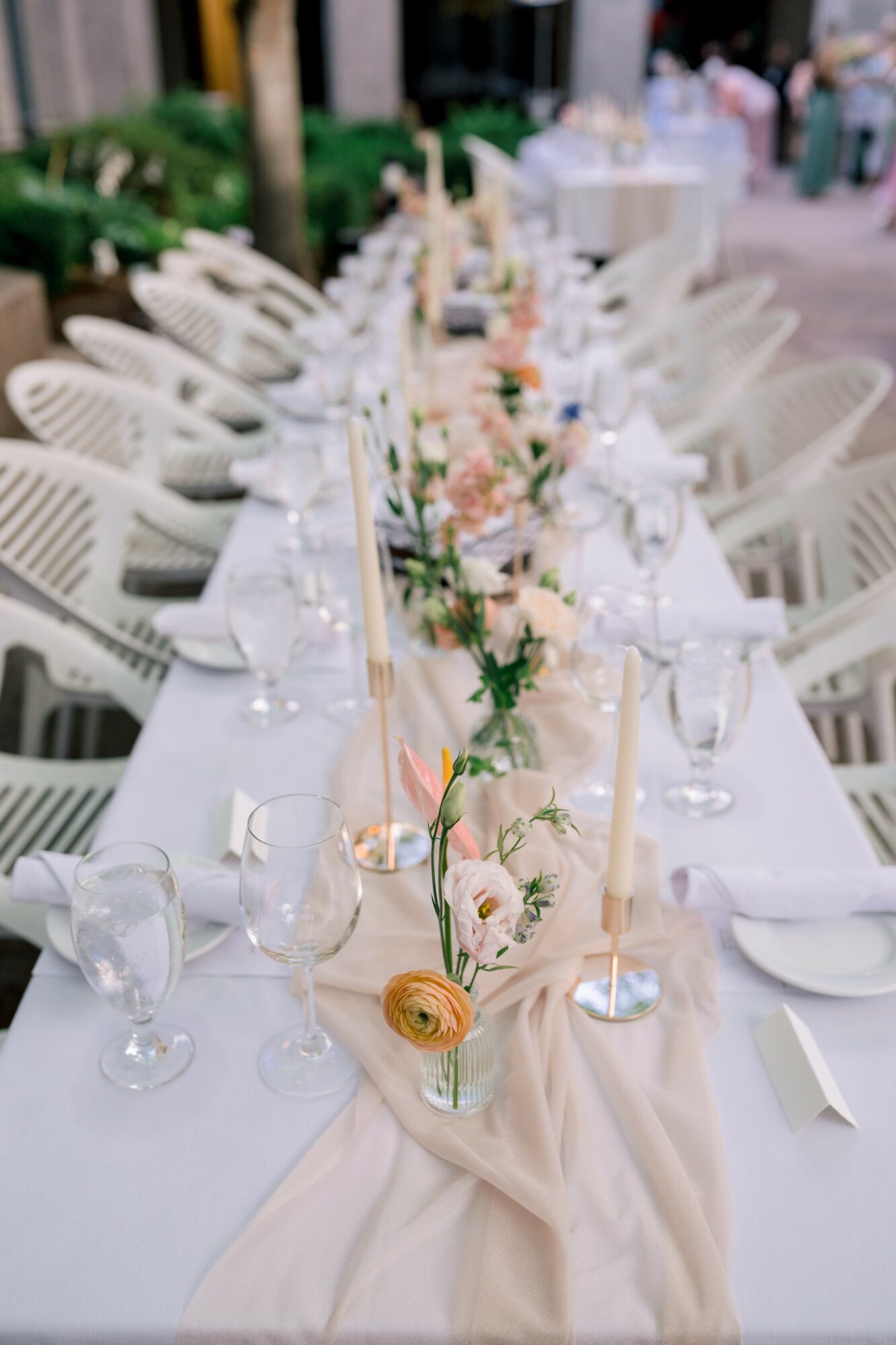 Long table set for an outdoor event with white chairs, floral centerpiece, candles, and tableware.