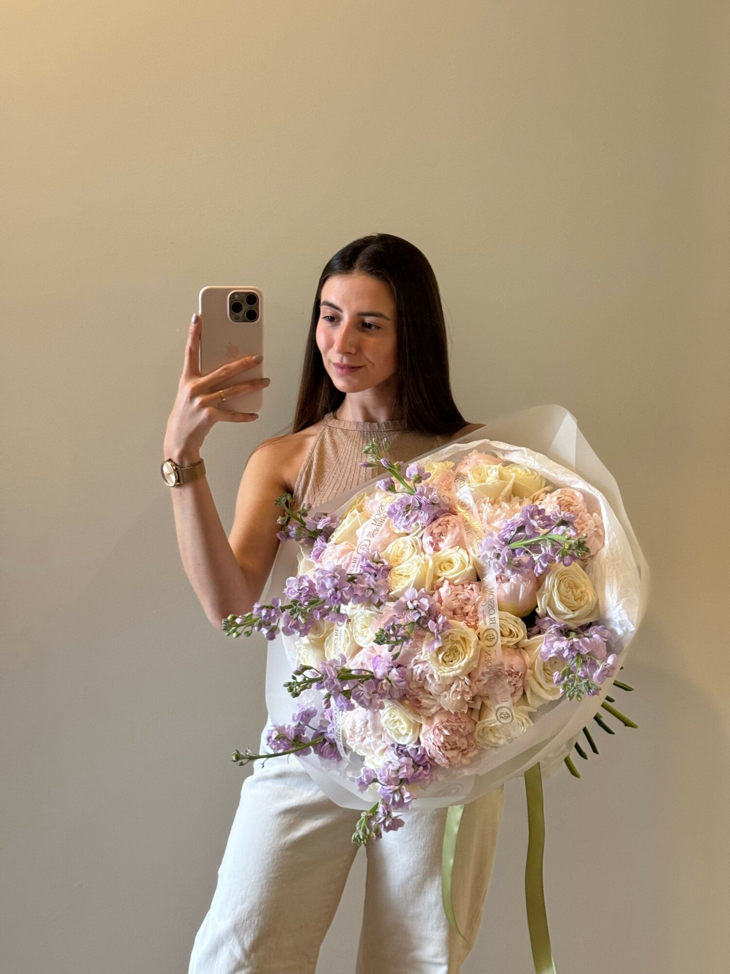 Woman holding a large bouquet of pastel-colored flowers, taking a selfie in front of a plain wall.