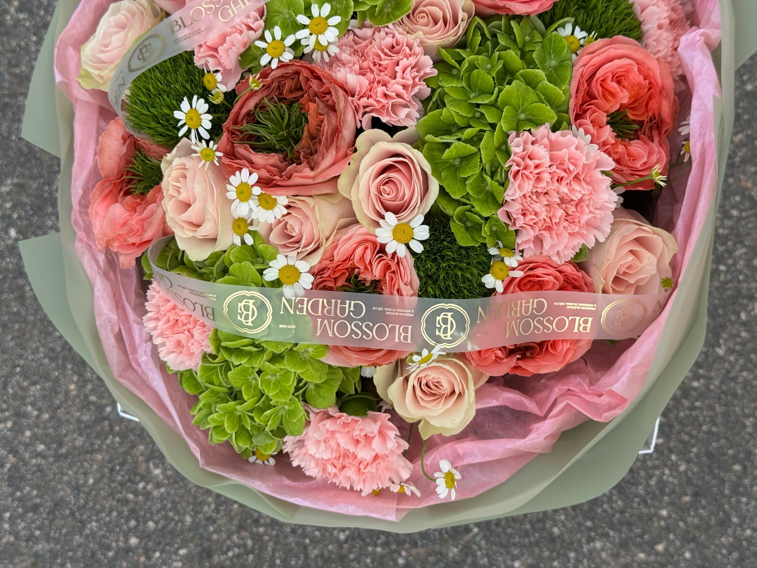 Bouquet of pink roses, green hydrangeas, white daisies, and pink carnations wrapped in pink paper.