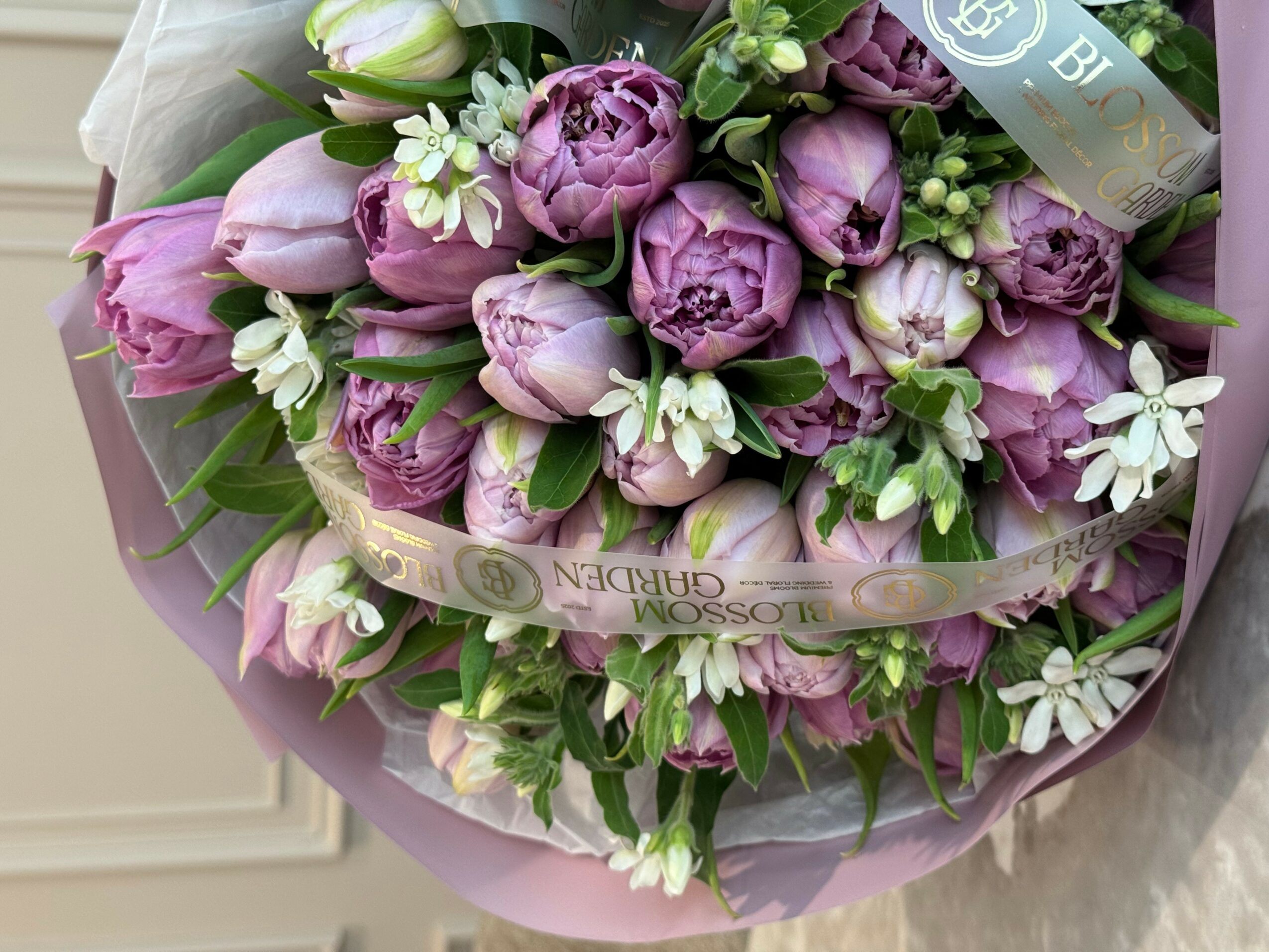 Bouquet of purple and white flowers with green leaves, wrapped in pink paper.