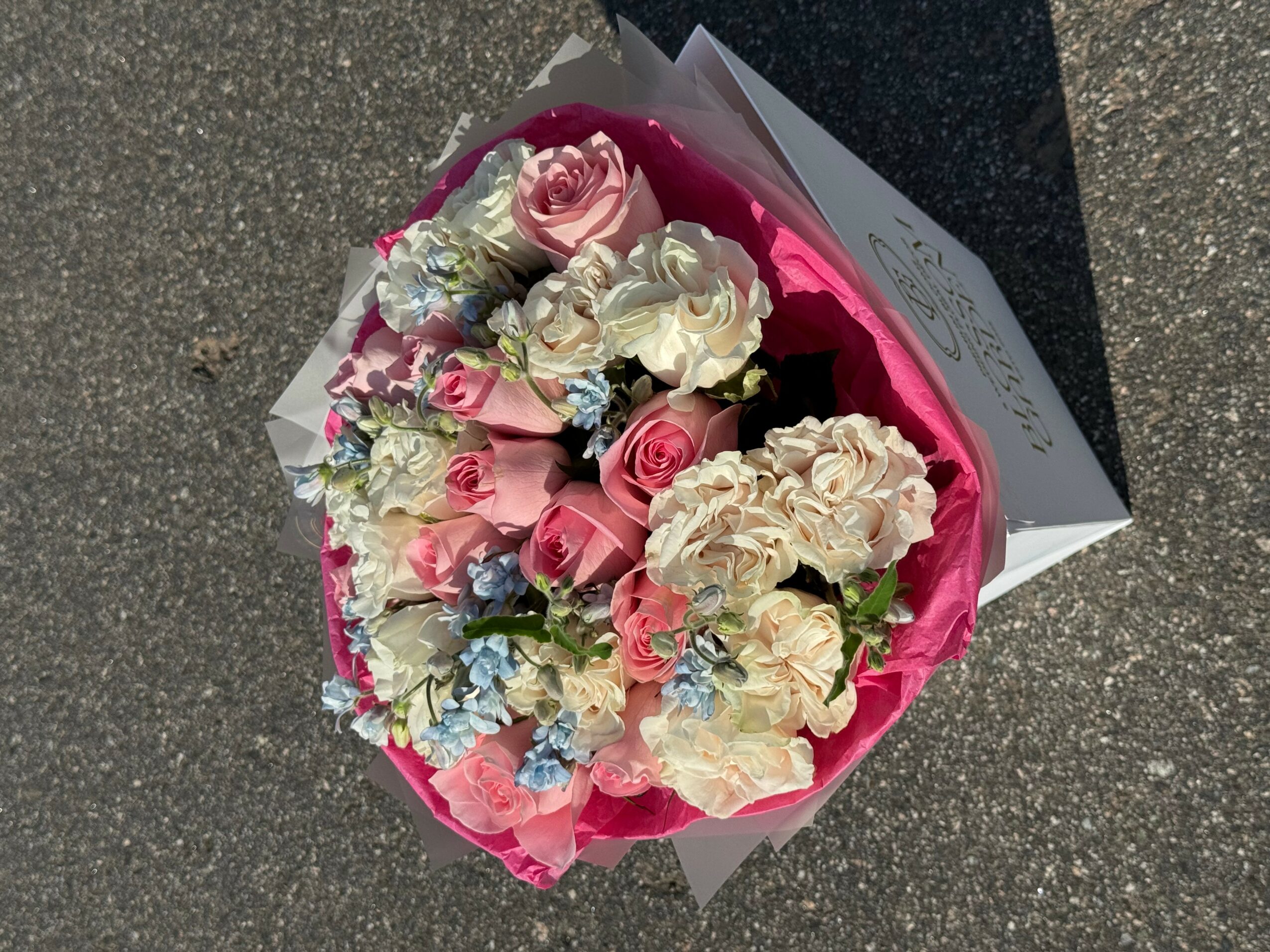 Bouquet of mixed pink, white, and blue flowers wrapped in pink paper, resting on a dark surface.