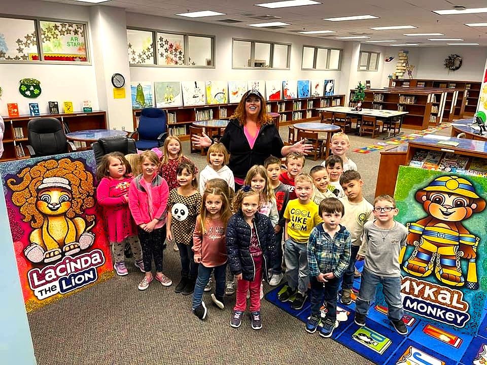 Group of children and a woman in a classroom with colorful posters and books, some children holding toys or objects.
