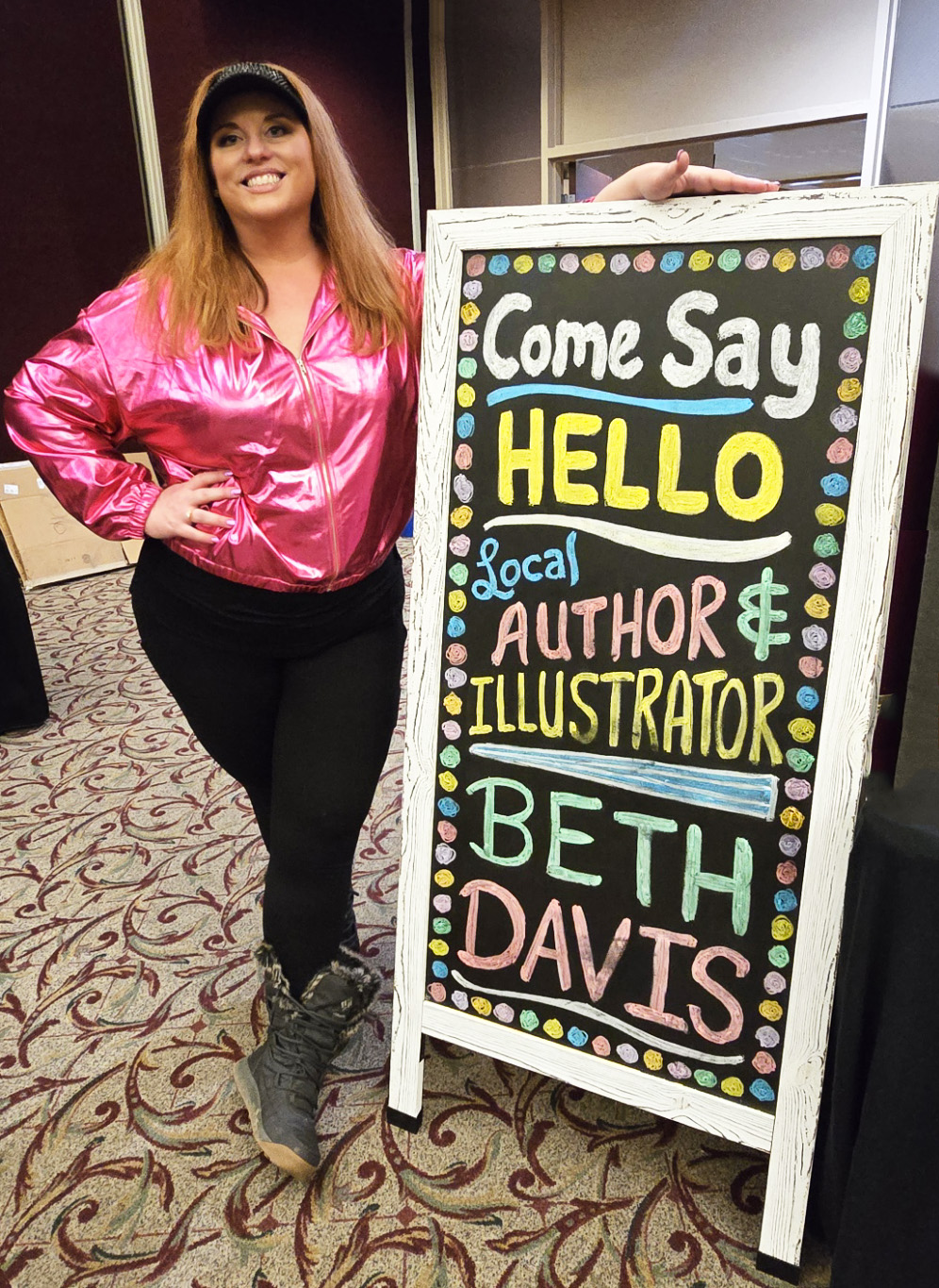 Woman in pink jacket standing next to a colorful signboard with text about an author and illustrator named Beth Davis.