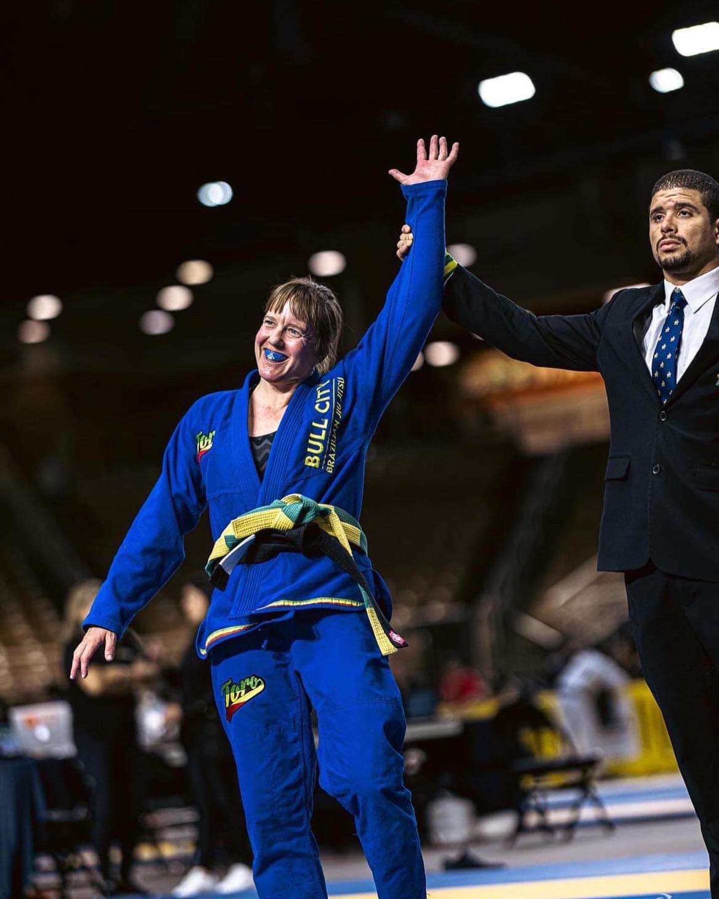 Female athlete in blue judo uniform smiling and raising her hand, with a male official holding her hand up, in an indoor sports arena.