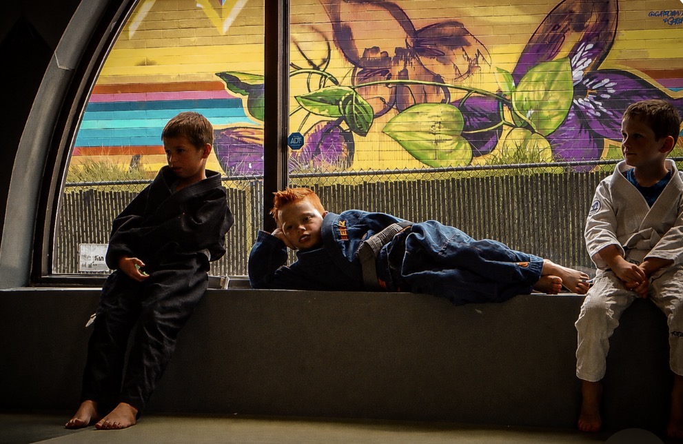 Three children sitting and lying on a bench in front of a colorful mural with flowers and leaves, indoors.