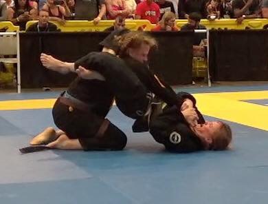 Two women practicing Brazilian Jiu-Jitsu on a blue mat, one on top of the other, in a competition setting.