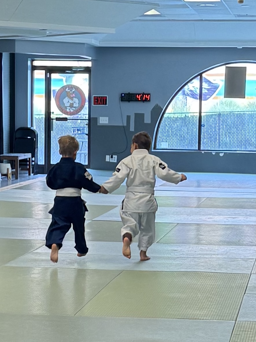 Two children in martial arts uniforms holding hands and walking on a mat in a training room.