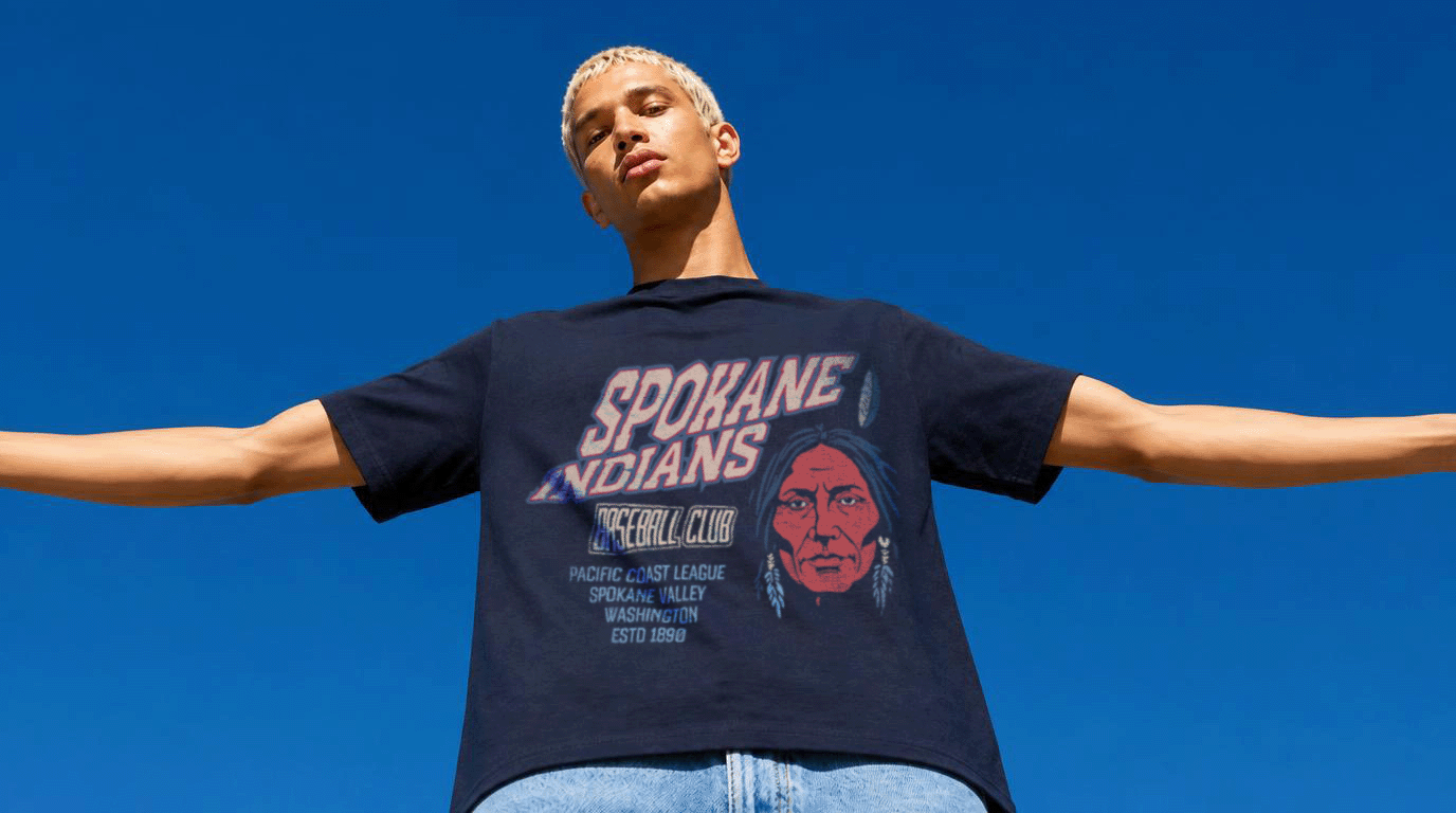 Person with arms outstretched wearing a graphic T-shirt against a blue sky.
