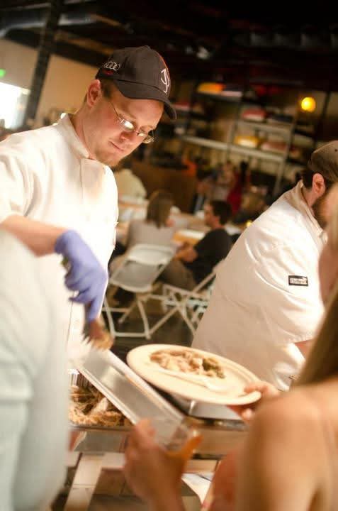 Person in white chef's coat and black cap serving food at a busy restaurant or event, with other staff and guests in background.