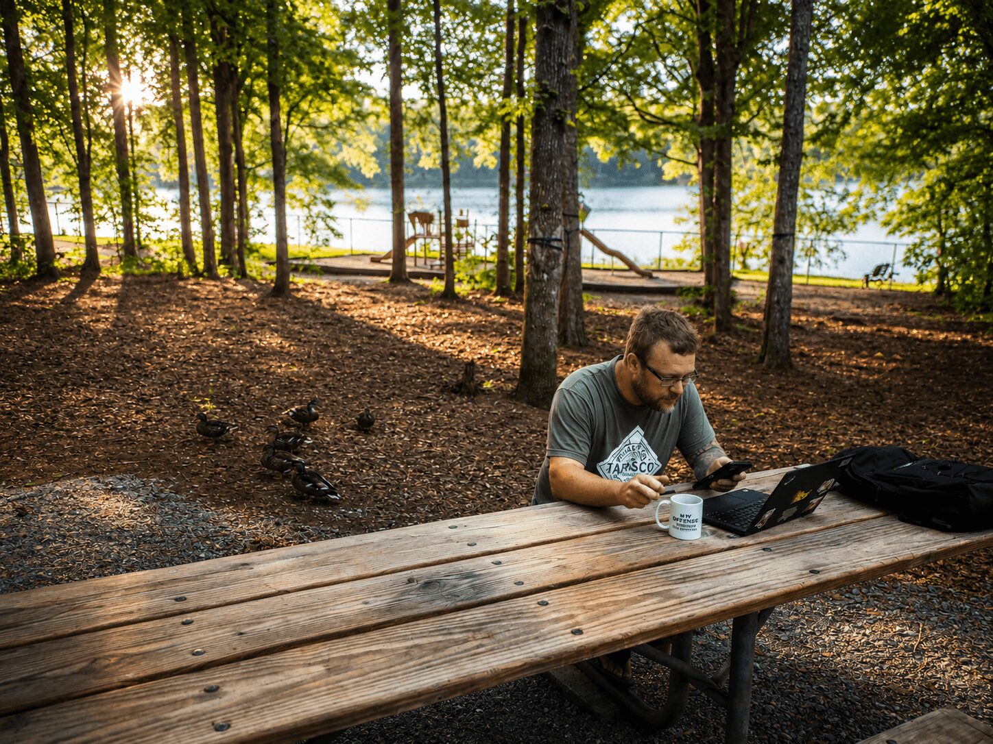 Man sitting at a picnic table in a wooded area near a lake, working on a laptop with trees and water in the background.