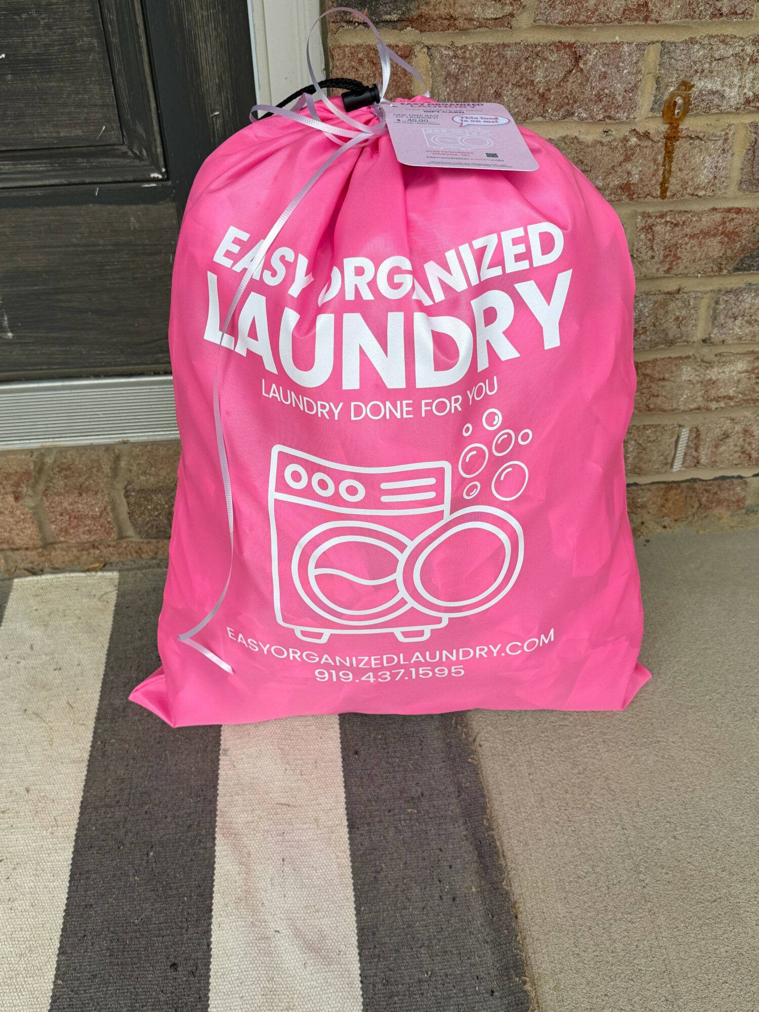 Pink laundry bag with white text and icons, placed on a sidewalk near a brick wall and a black door.
