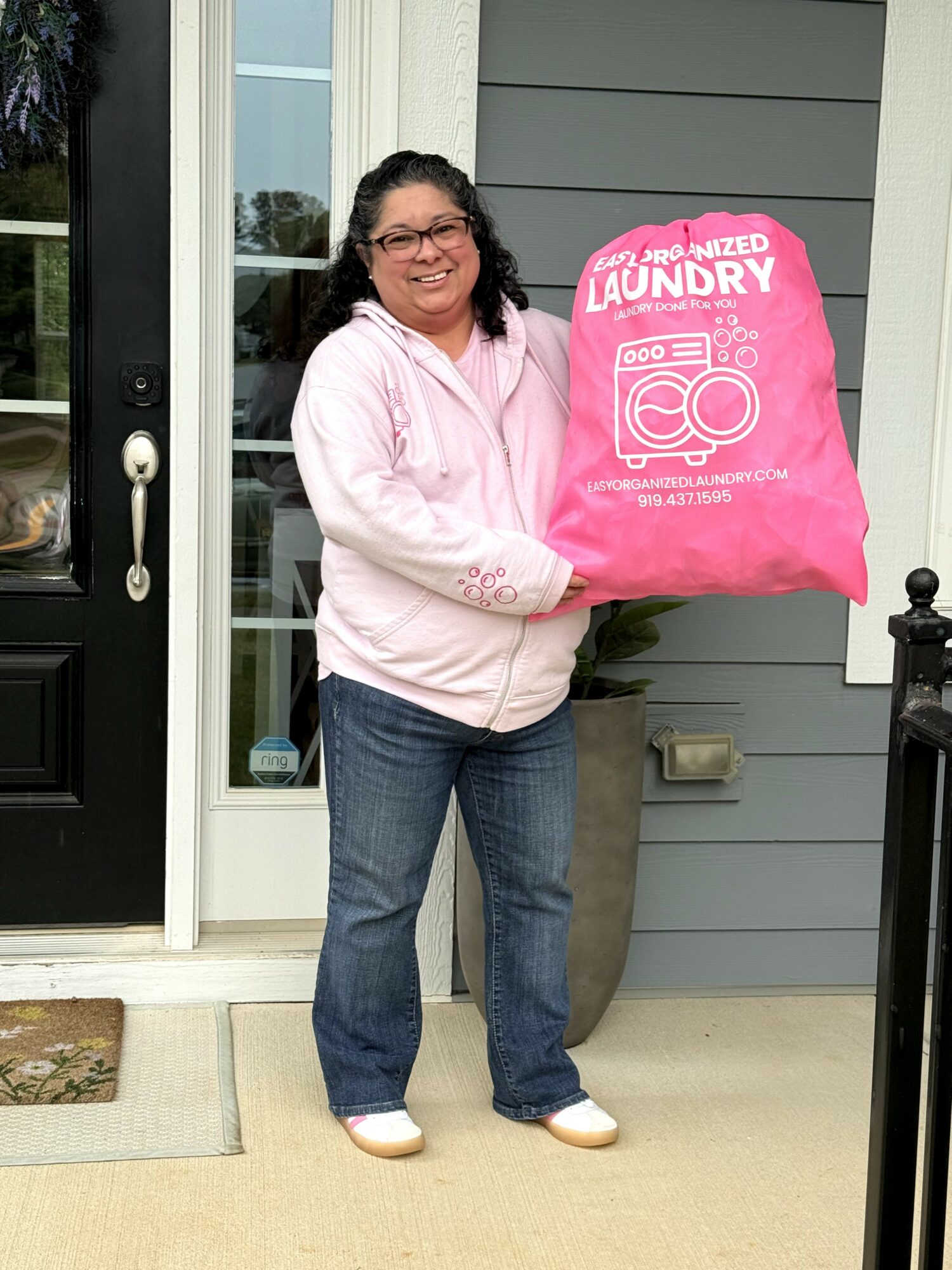 Woman holding a pink laundry bag outside a house, smiling, wearing glasses, pink hoodie, jeans, and white shoes.