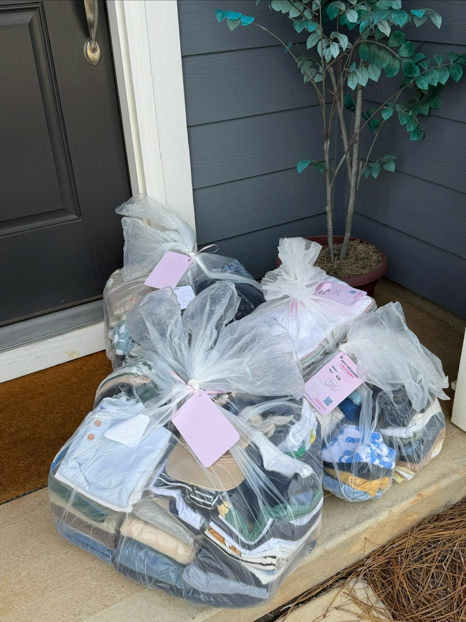 Four large transparent bags filled with clothing and shoes are placed outside a door on a porch, with a potted plant nearby.