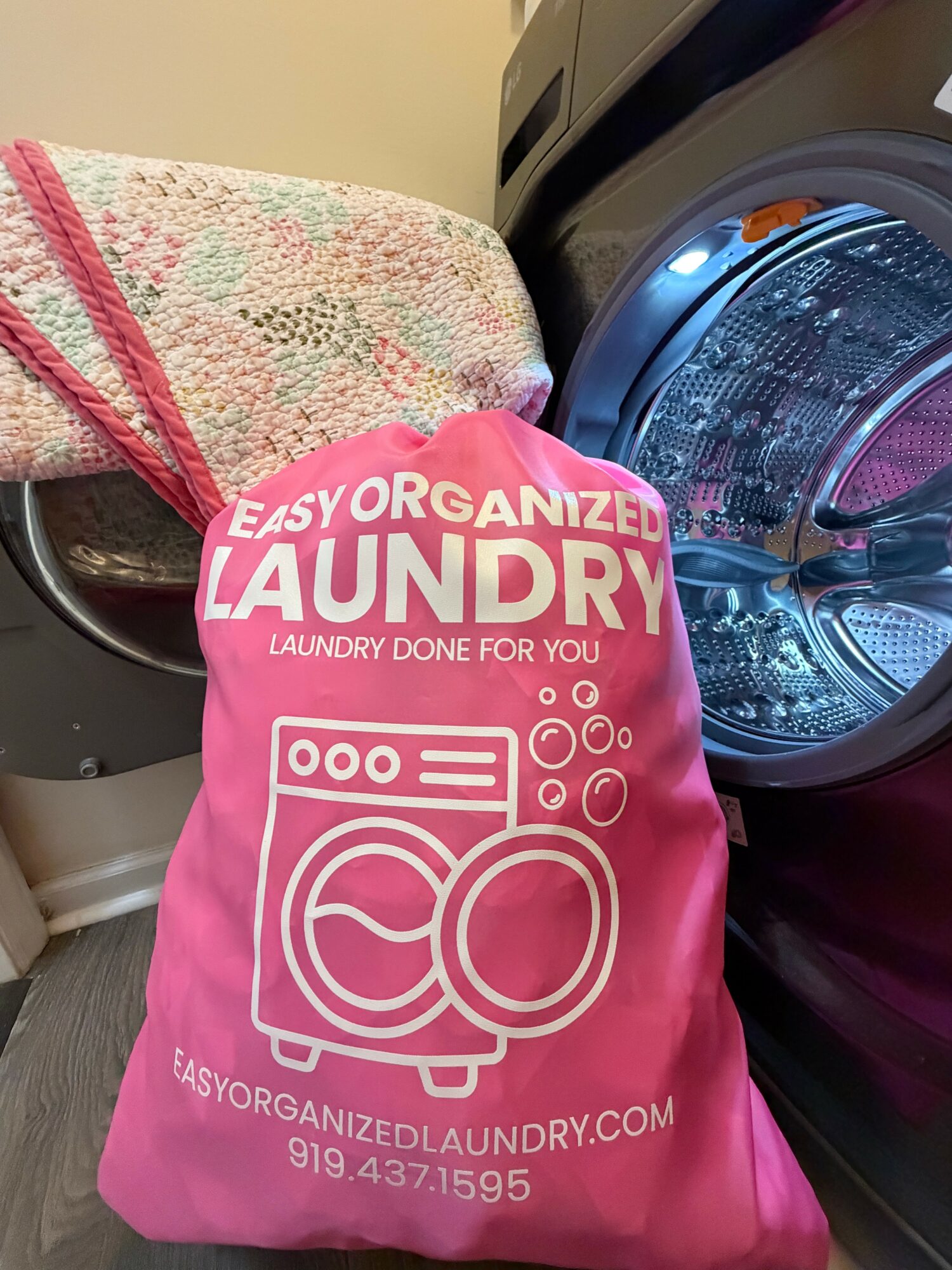 Pink laundry bag with white text and logo, placed in front of a washing machine, laundry basket on top, in a laundry room.