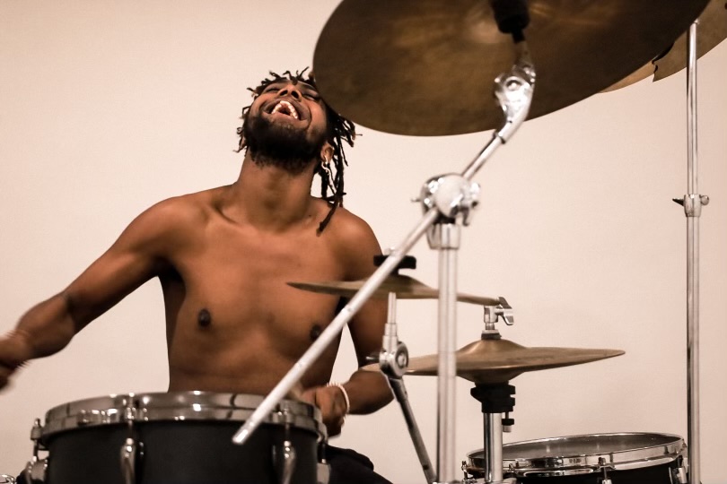 A shirtless man with dreadlocks plays drums, looking upward with a joyful expression, against a plain background.